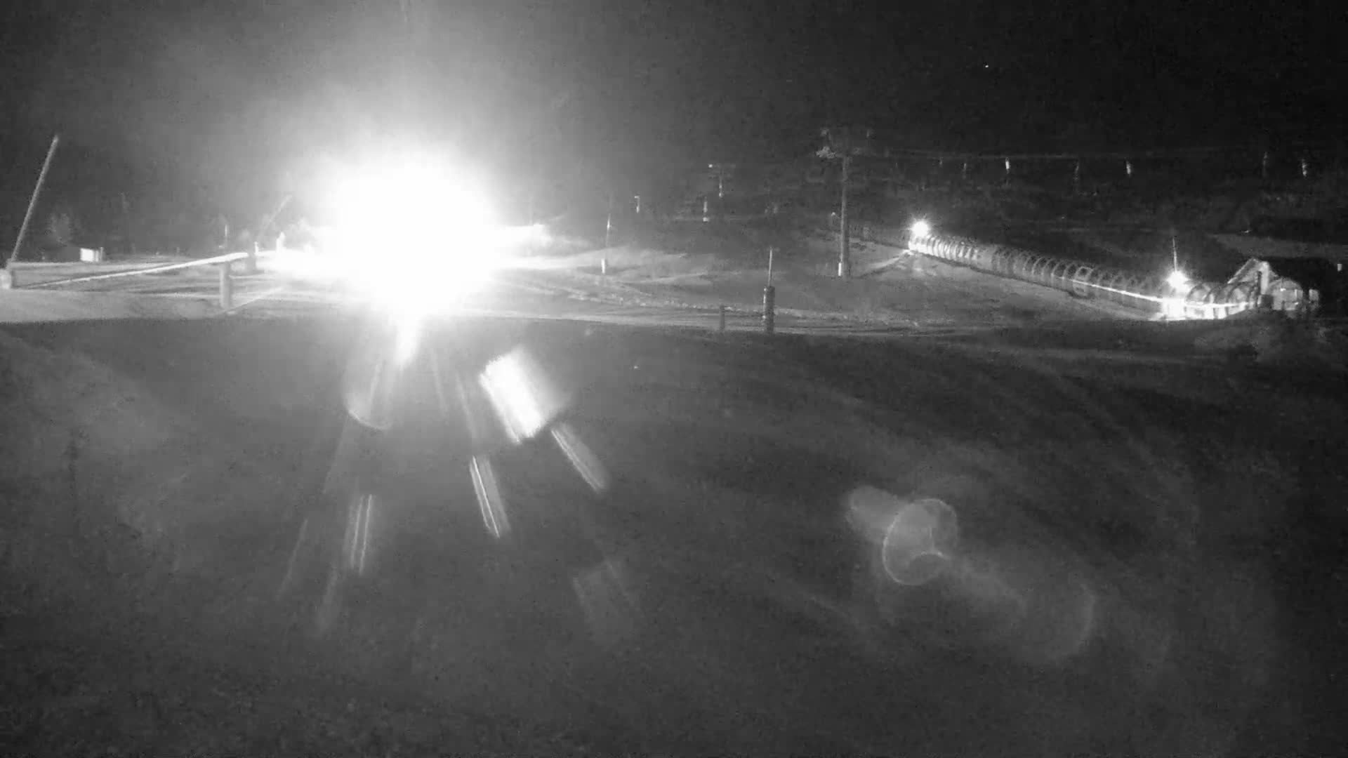 A snow-covered ski slope featuring several red marker poles, a distant ski lift with empty chairs, and a building obscured by dense fog or whiteout conditions.