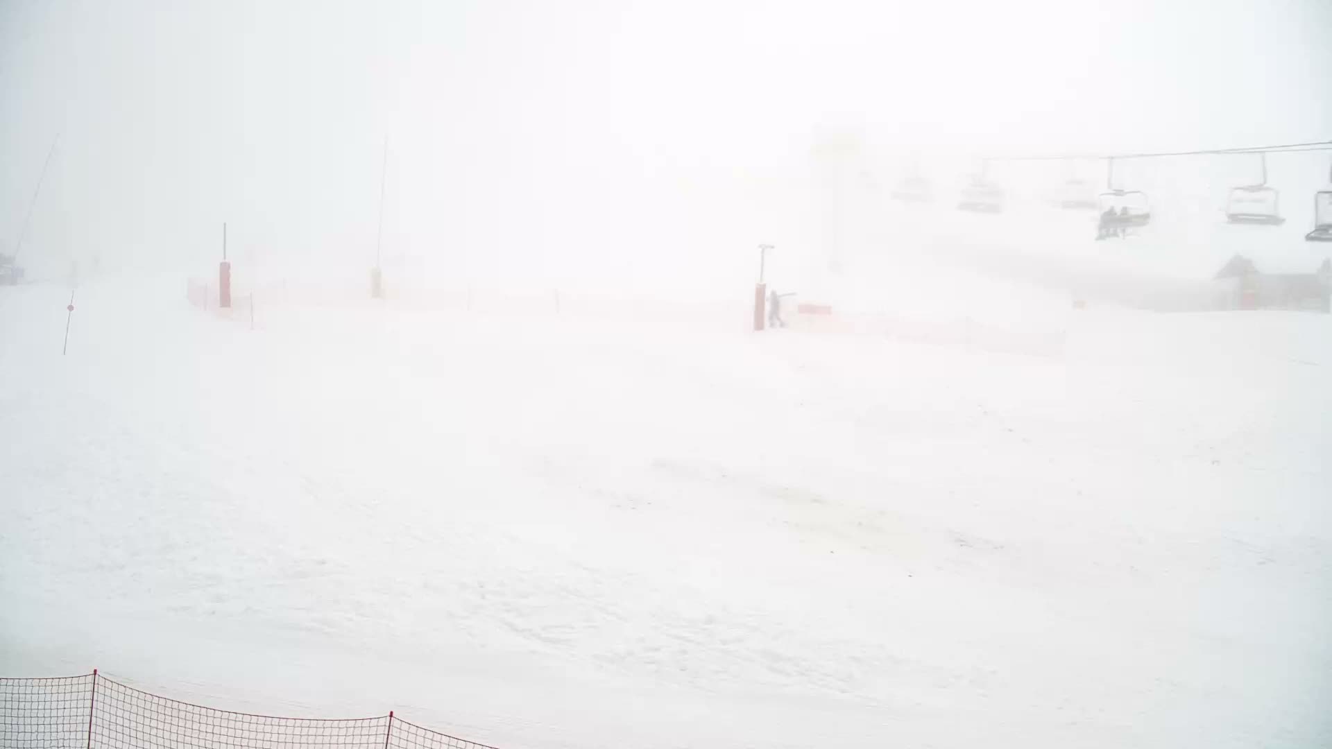 A snow-covered ski slope featuring several red marker poles, a distant ski lift with empty chairs, and a building obscured by dense fog or whiteout conditions.