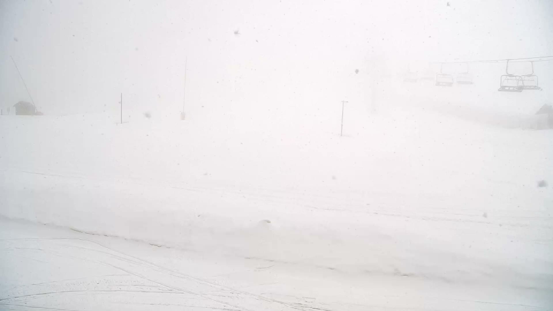 A snow-covered ski slope featuring several red marker poles, a distant ski lift with empty chairs, and a building obscured by dense fog or whiteout conditions.