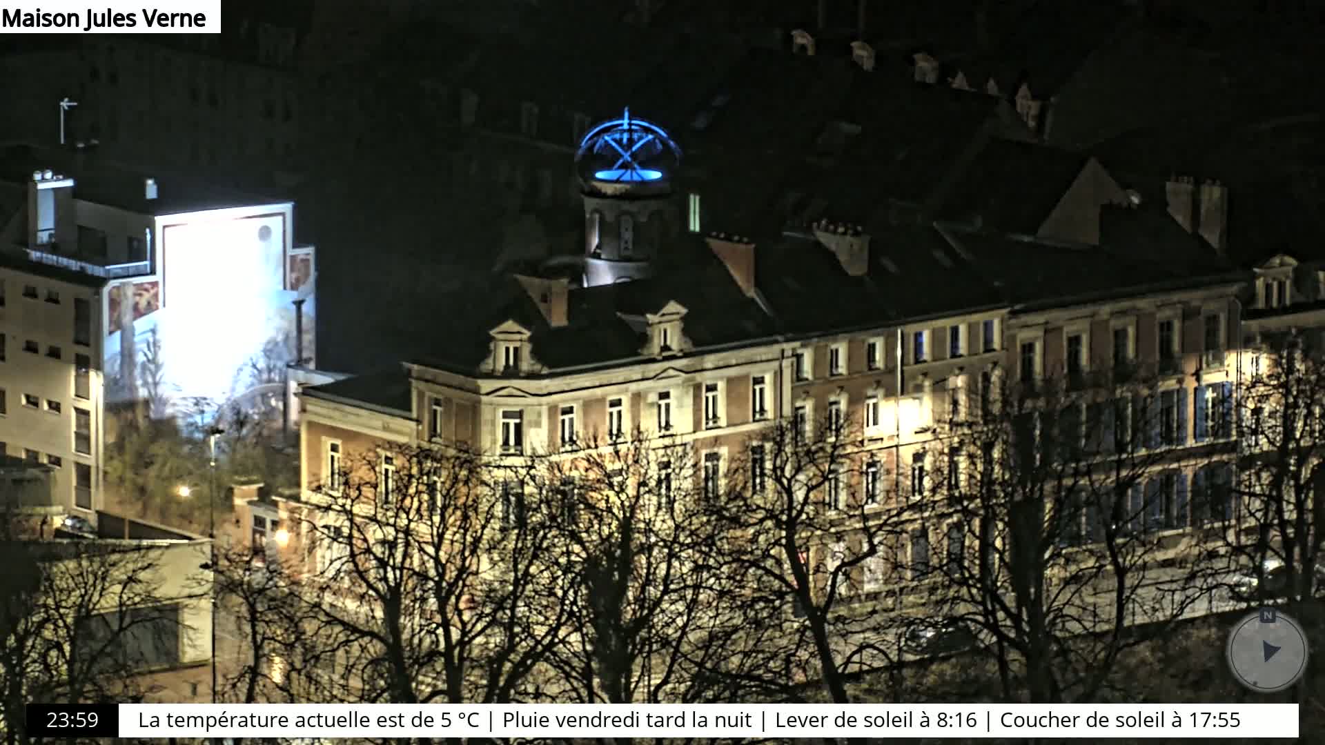 A nighttime view of a large building with a blue-lit dome, surrounded by bare trees under rainy conditions.