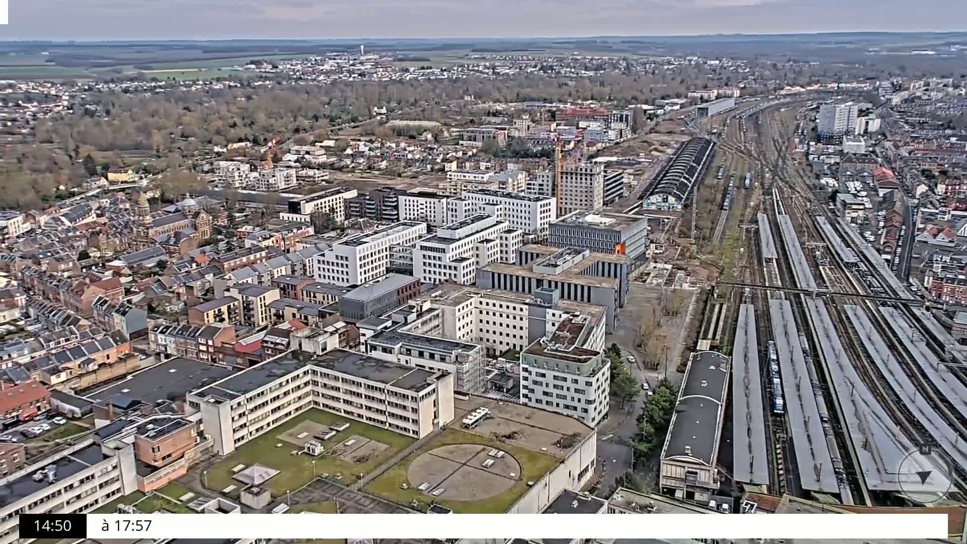 An aerial view shows a city with a mix of residential and commercial buildings, including a large hospital complex, near a sprawling railway yard under an overcast sky.
