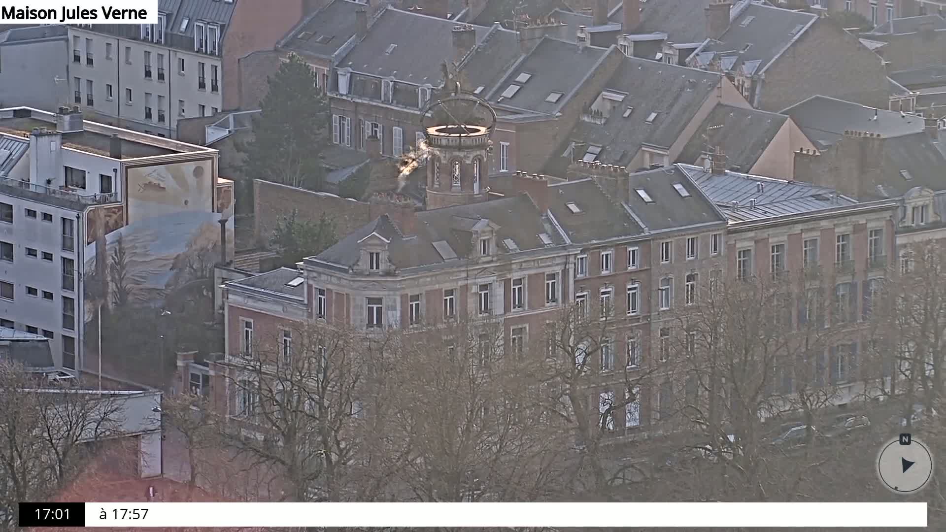 An aerial view shows a city block of buildings with brown rooftops and bare trees under an overcast sky.