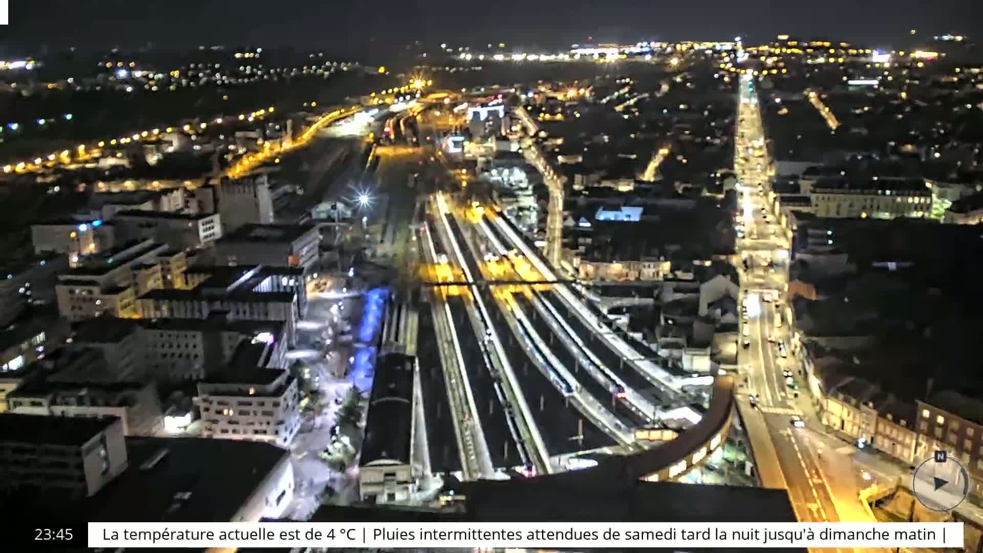A nighttime aerial view of a city's illuminated train station and surrounding streets, with intermittent rain expected.