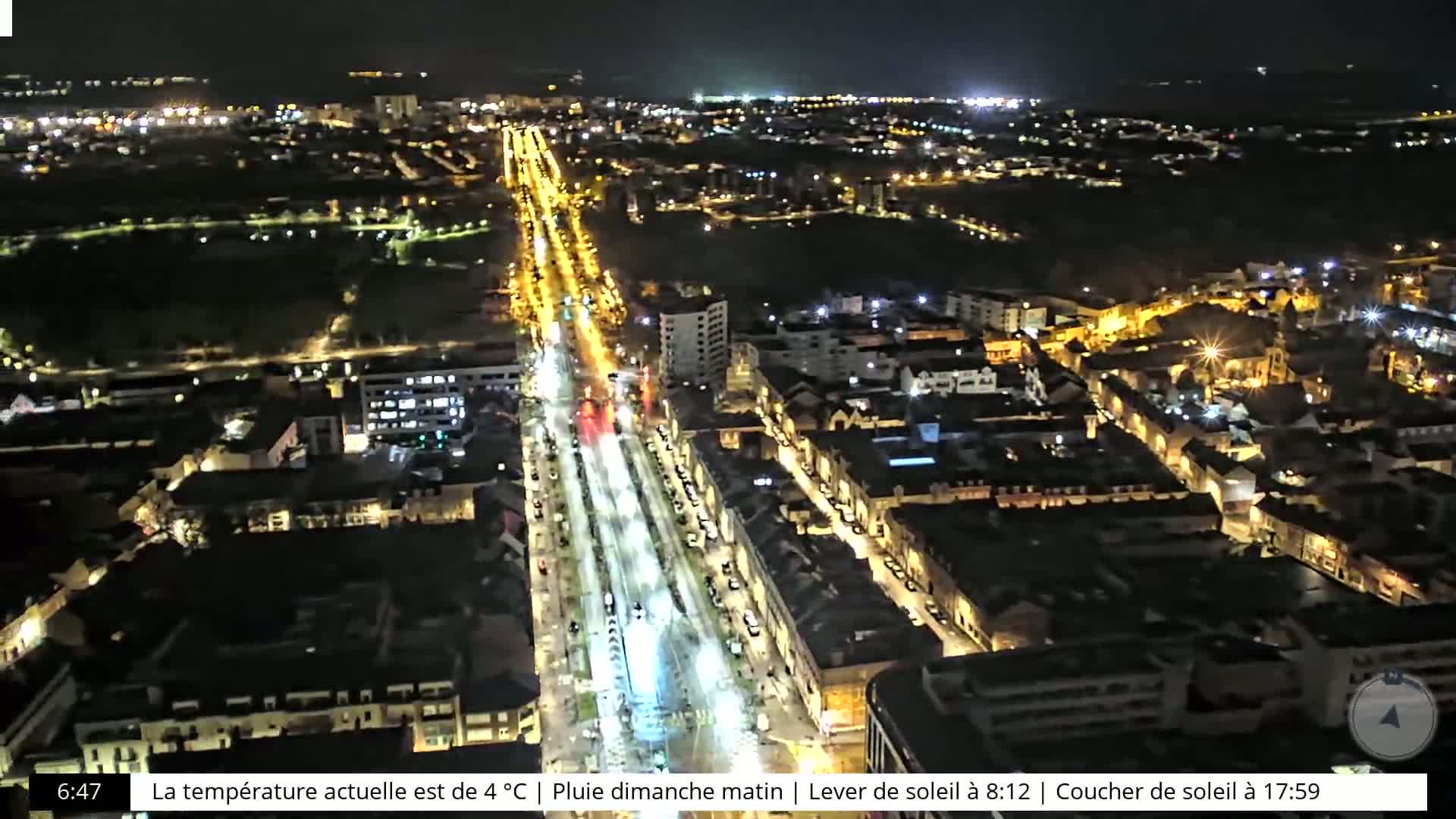 A nighttime aerial view of a city with brightly lit streets and buildings, under rainy conditions.