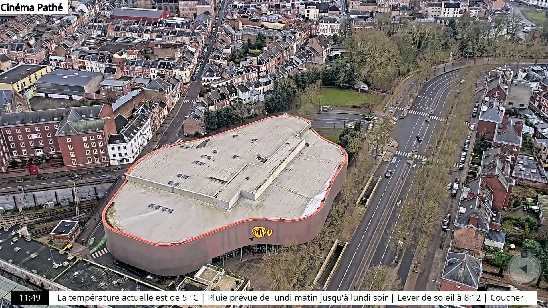 An aerial view shows a curved-roof building, possibly a cinema, situated amongst residential buildings and roads, with light traffic and bare winter trees under overcast skies.