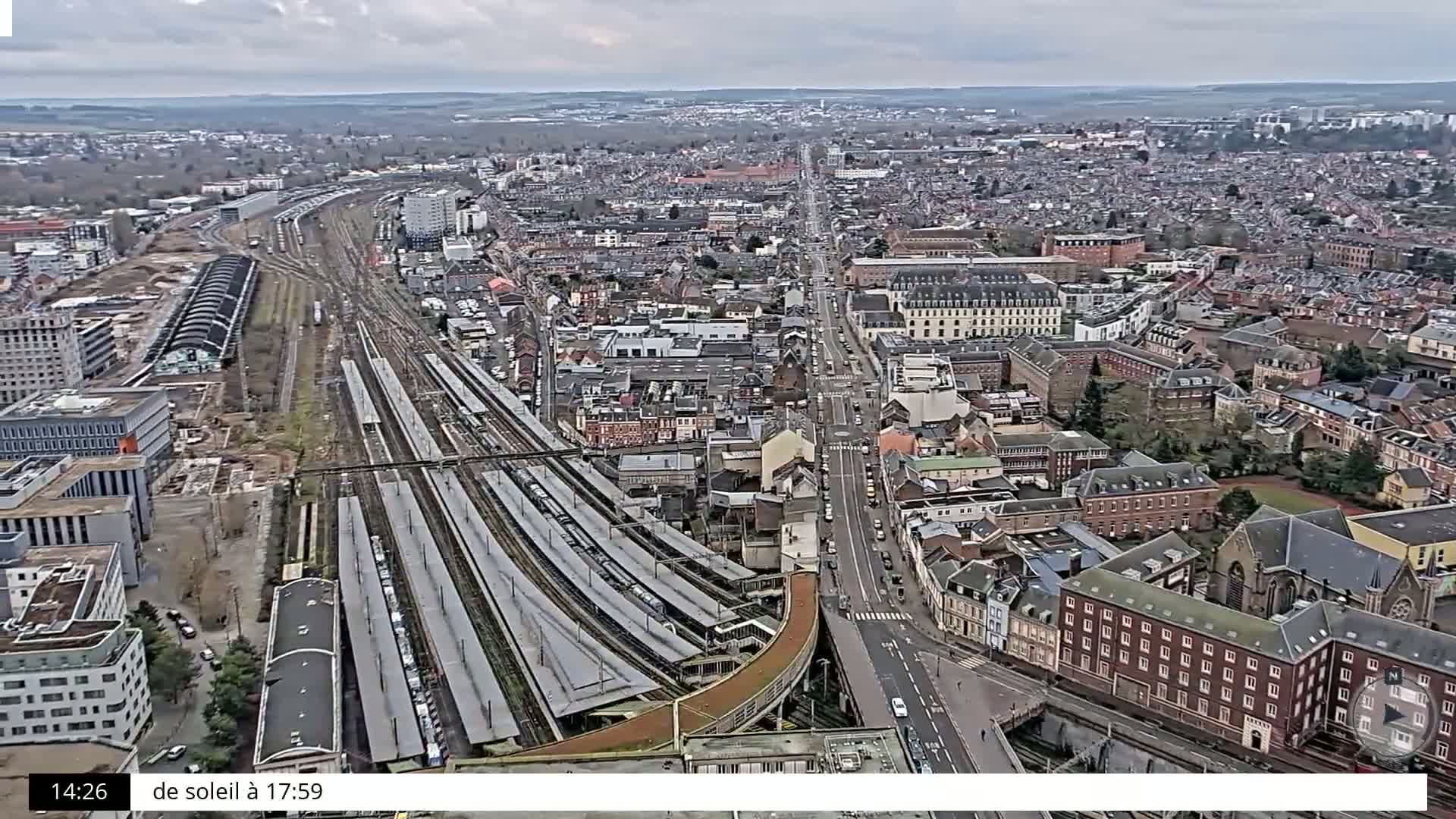 An aerial view shows a city with a large train station, many buildings, and a main road running through it on an overcast day.