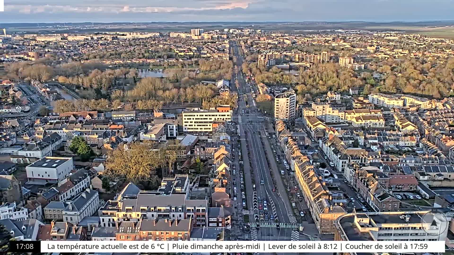An aerial view of a city on an overcast day shows a long, straight road lined with parked cars and buildings, with a waterway visible in the background.