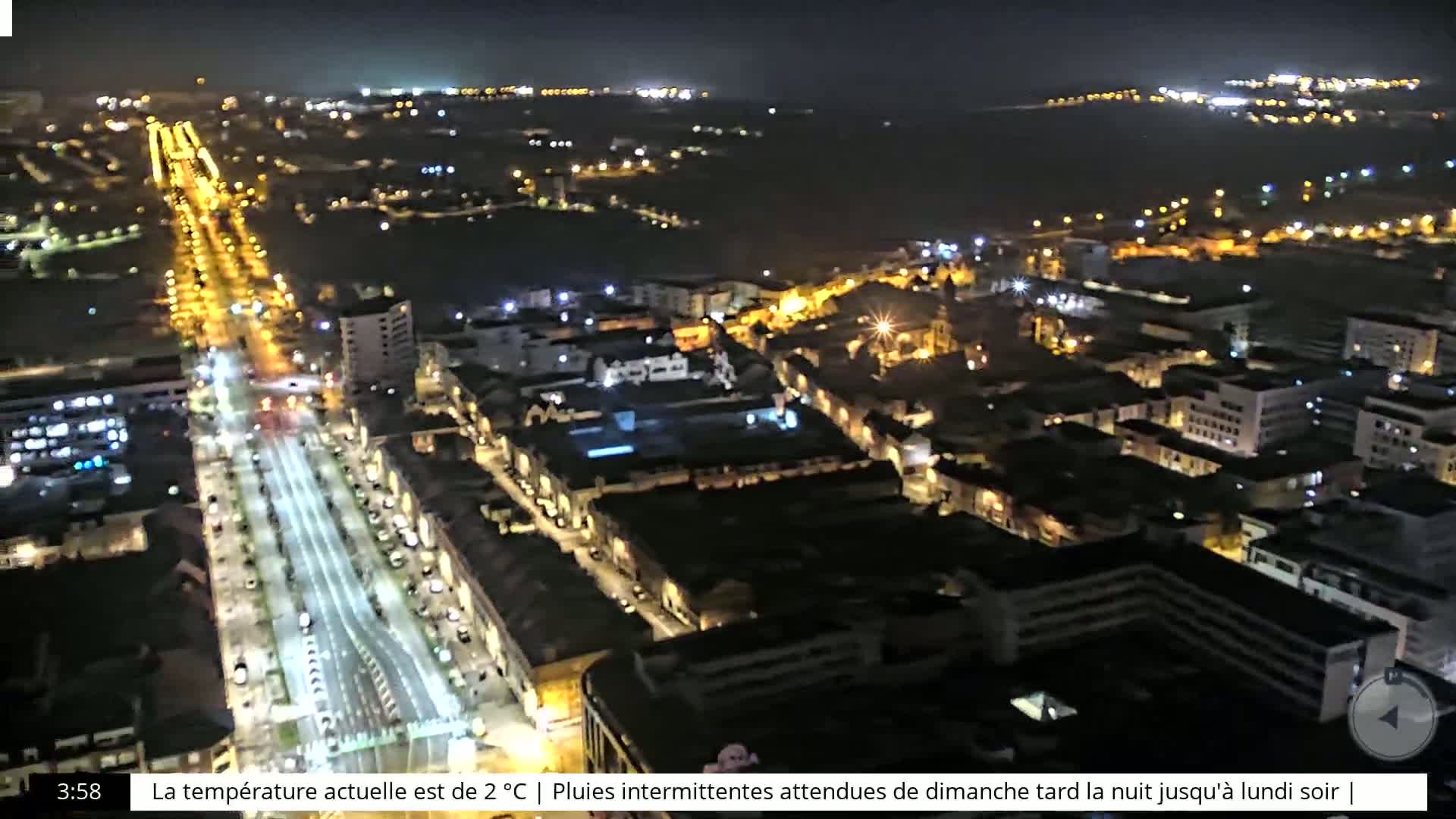 An aerial nighttime view of a city with brightly lit streets and buildings, extending to a dark body of water in the distance.