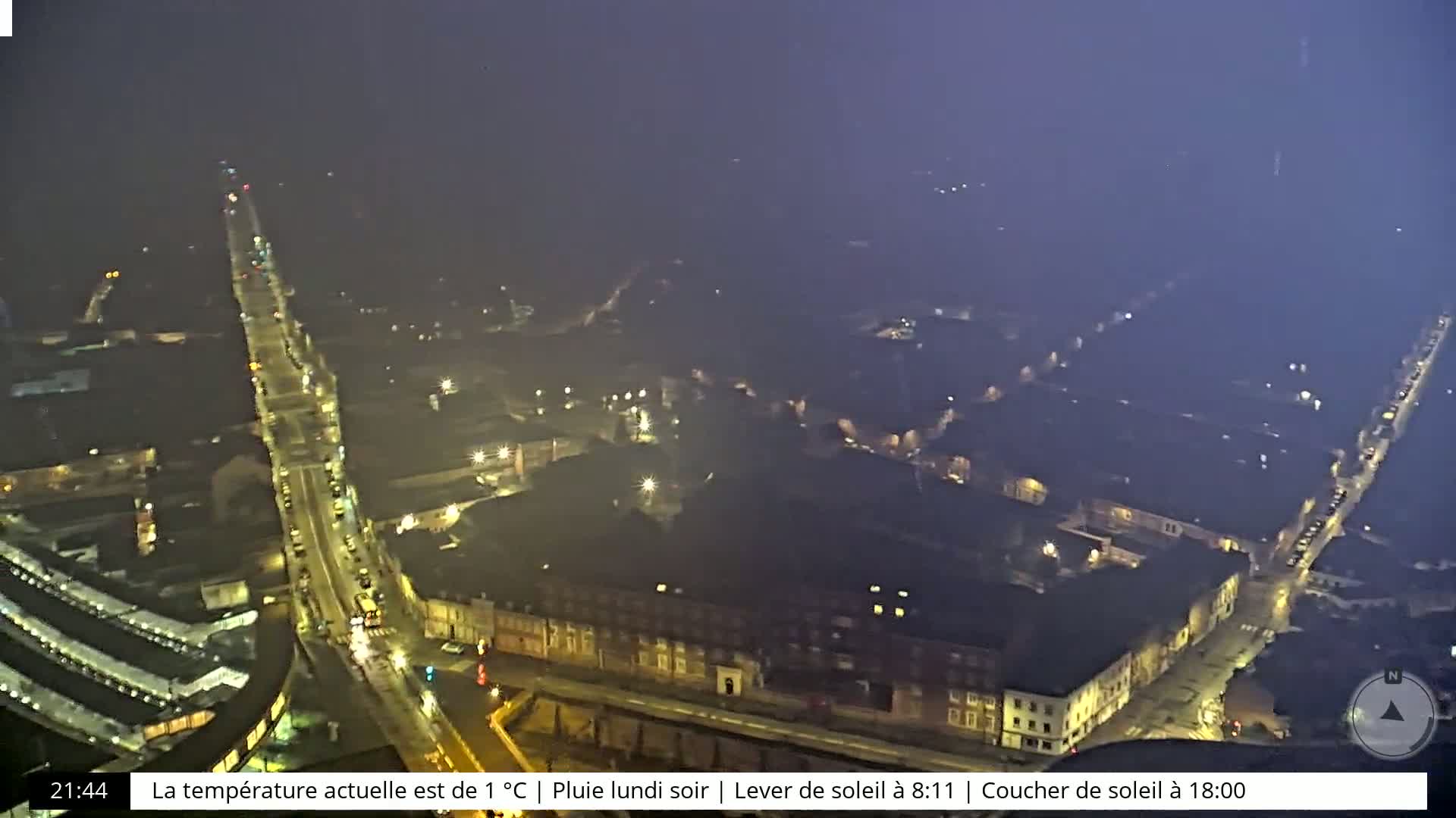 A nighttime, rainy view of a city street with dimly lit buildings and sparsely trafficked roads.