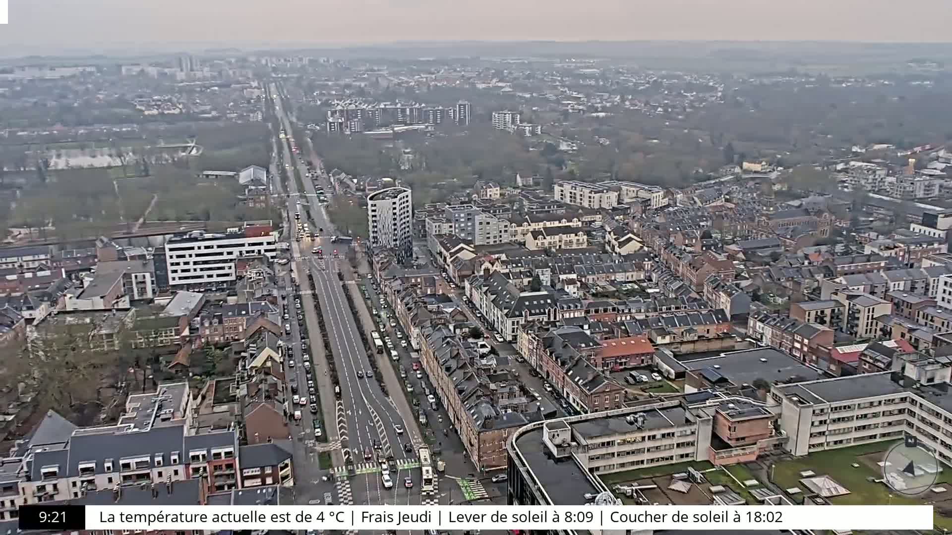 An aerial view of a city on an overcast day, showing a long road with traffic flanked by densely packed residential buildings.