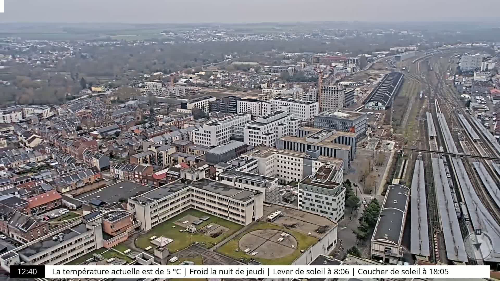 An aerial view of a city with many buildings, a large train station, and surrounding woods under an overcast sky.