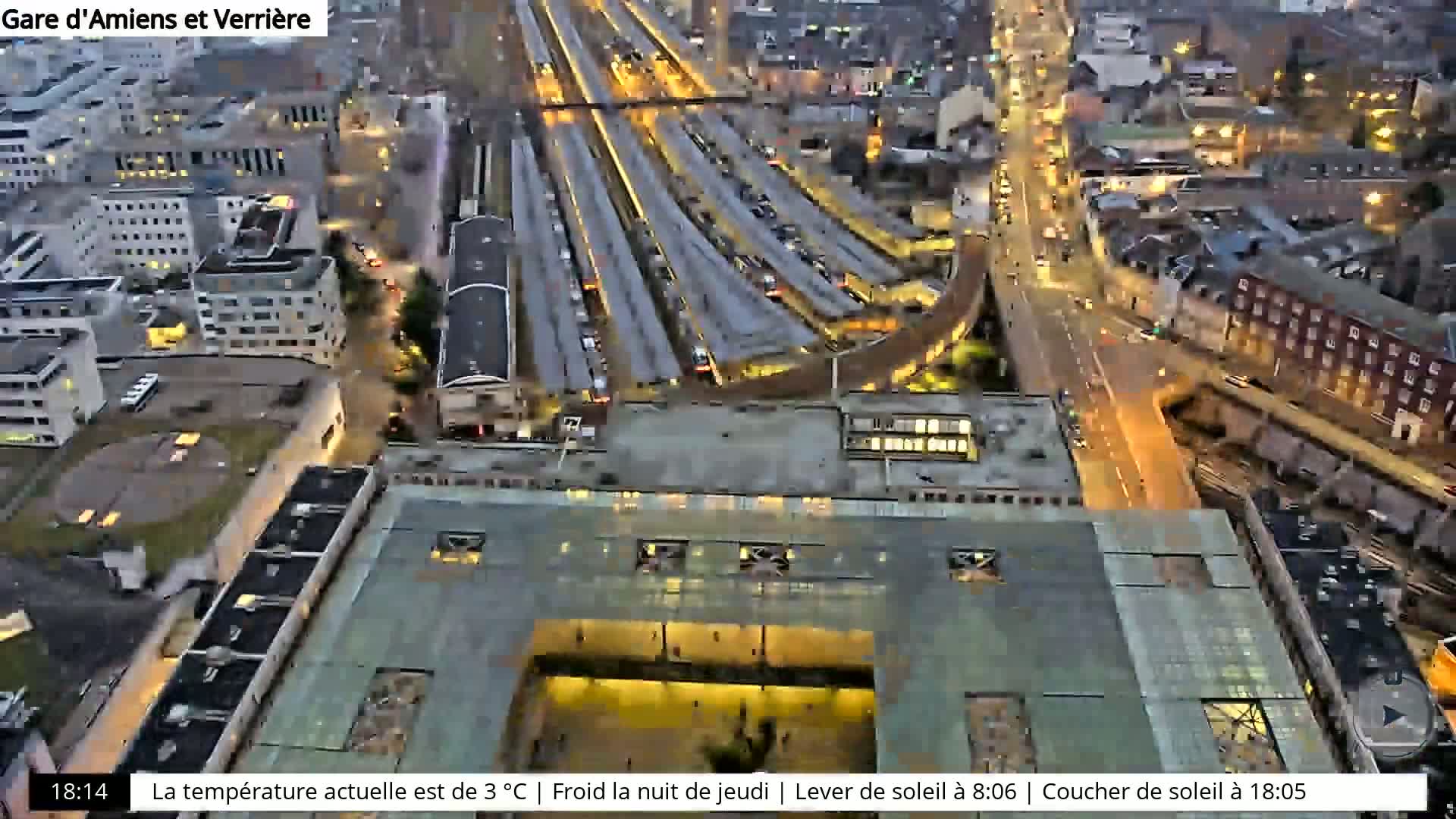 An aerial nighttime view of a city train station and surrounding buildings, with illuminated streets and a cool temperature.