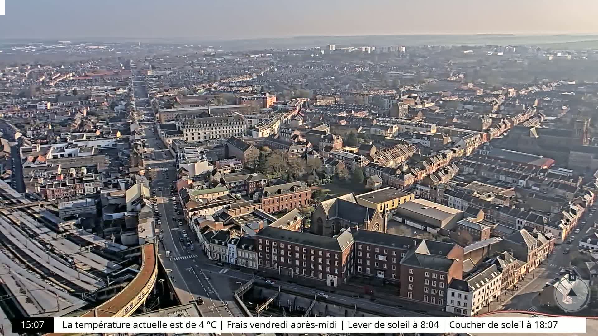 A high-angle, long shot of a city on a sunny, cool afternoon, showing a large road cutting through a dense area of buildings, with a railway line visible in the bottom-left corner.