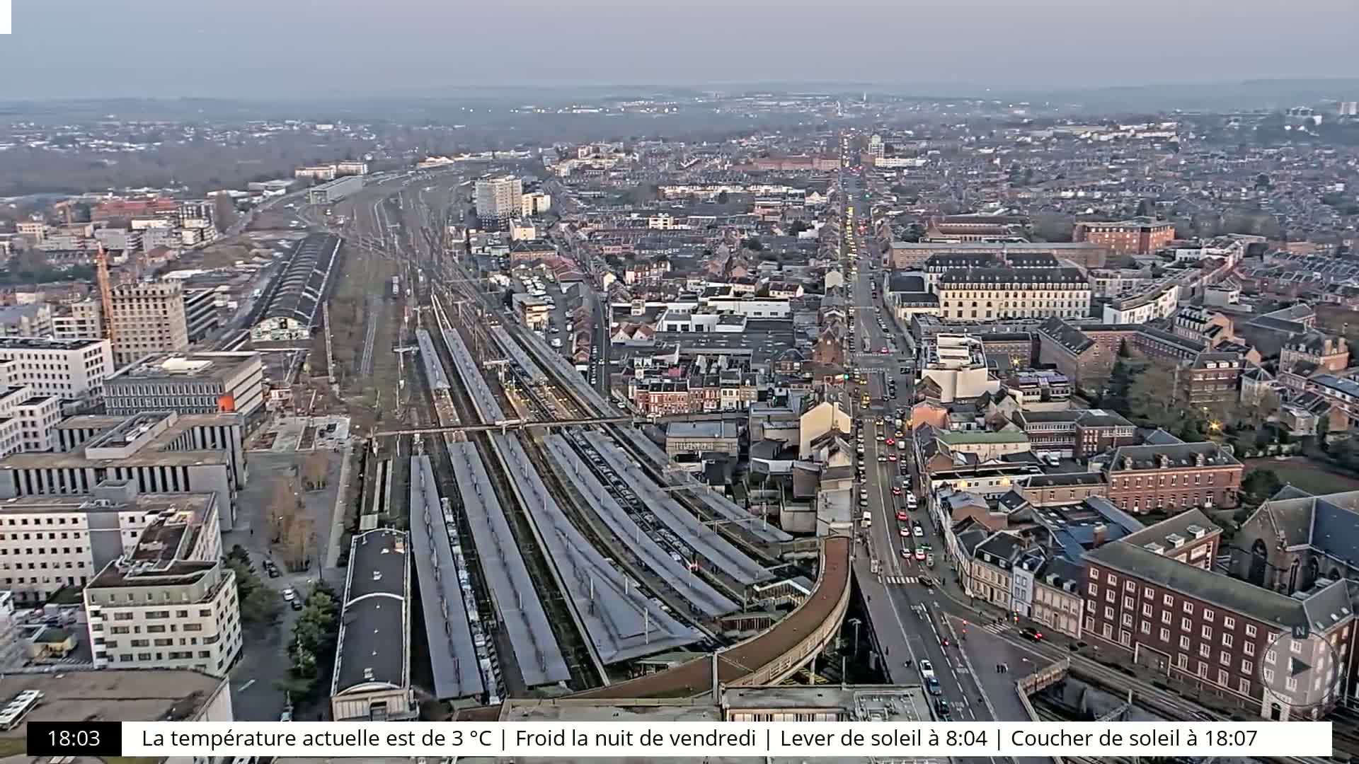 An aerial view shows a city with a train station and surrounding buildings under a twilight sky.
