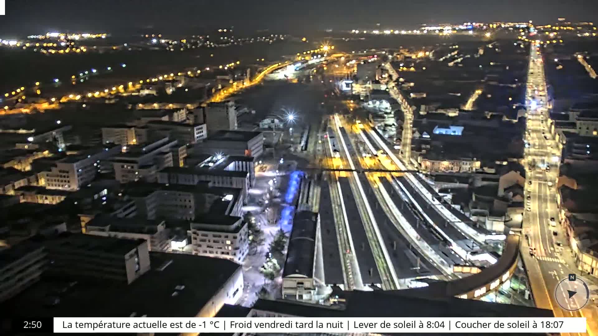 A nighttime aerial view of a city's illuminated train station and surrounding buildings, streets, and roadways.