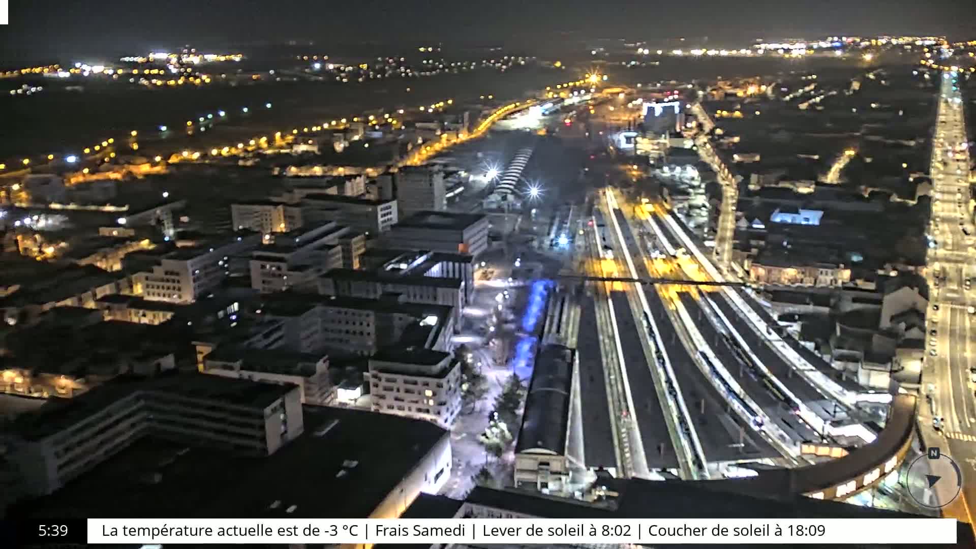 A nighttime, aerial view of a city's illuminated train station and surrounding buildings, under a clear sky.
