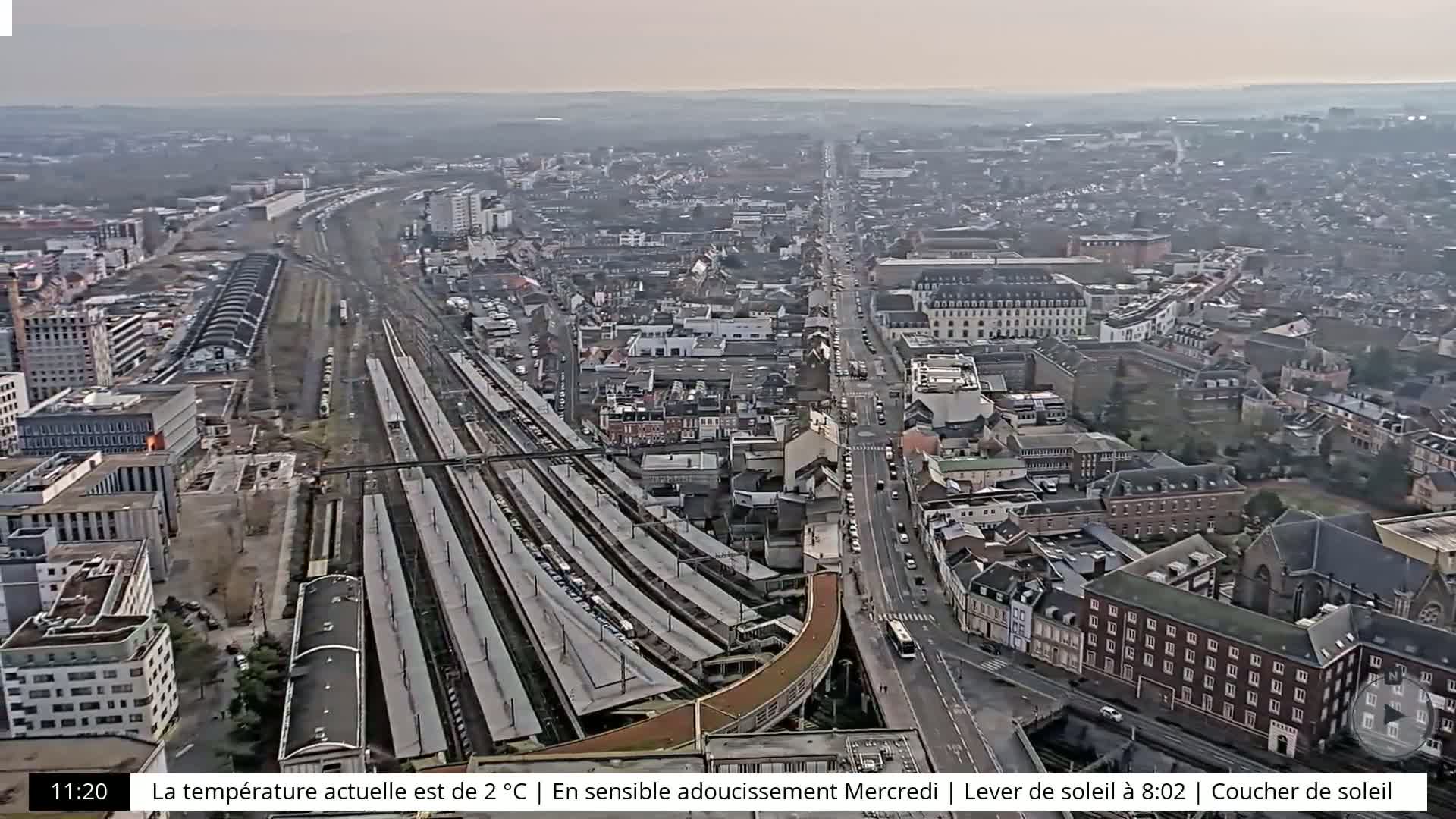 An aerial view shows a city with a train station and multiple tracks, under an overcast sky.