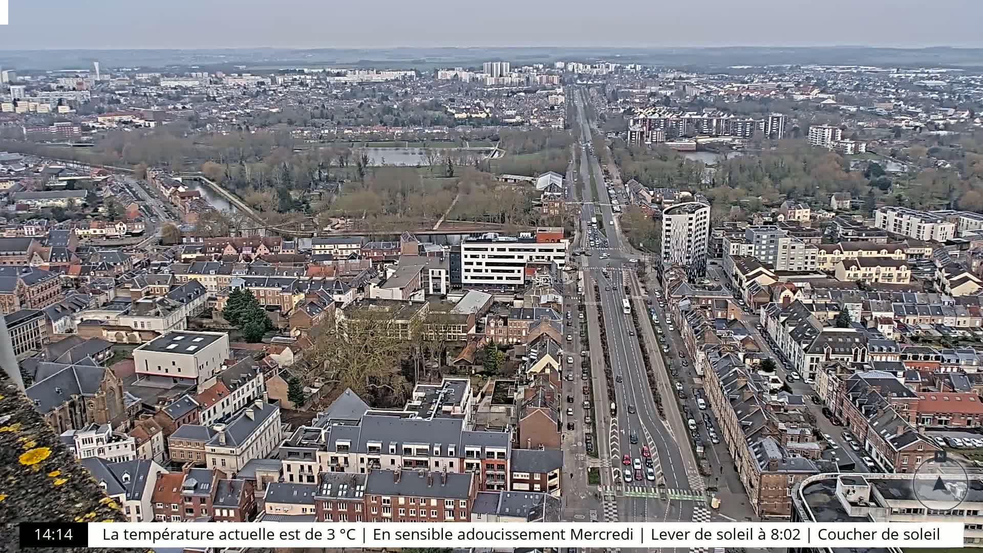 A high-angle, long shot of a city on a partly cloudy day shows a long road cutting through a densely packed area of low-rise buildings.