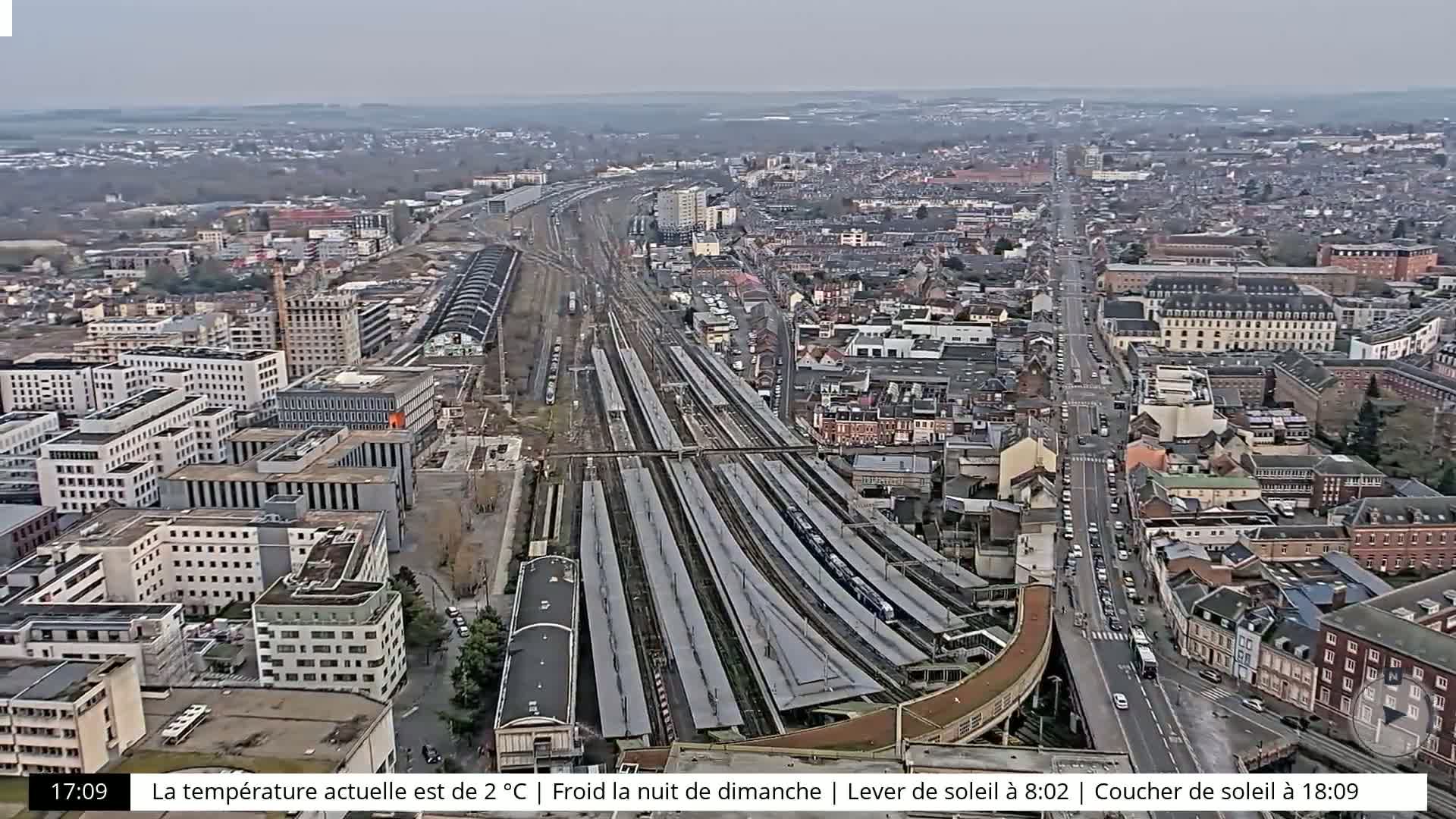 A high-angle, long shot of a city with a large train station, under an overcast sky.
