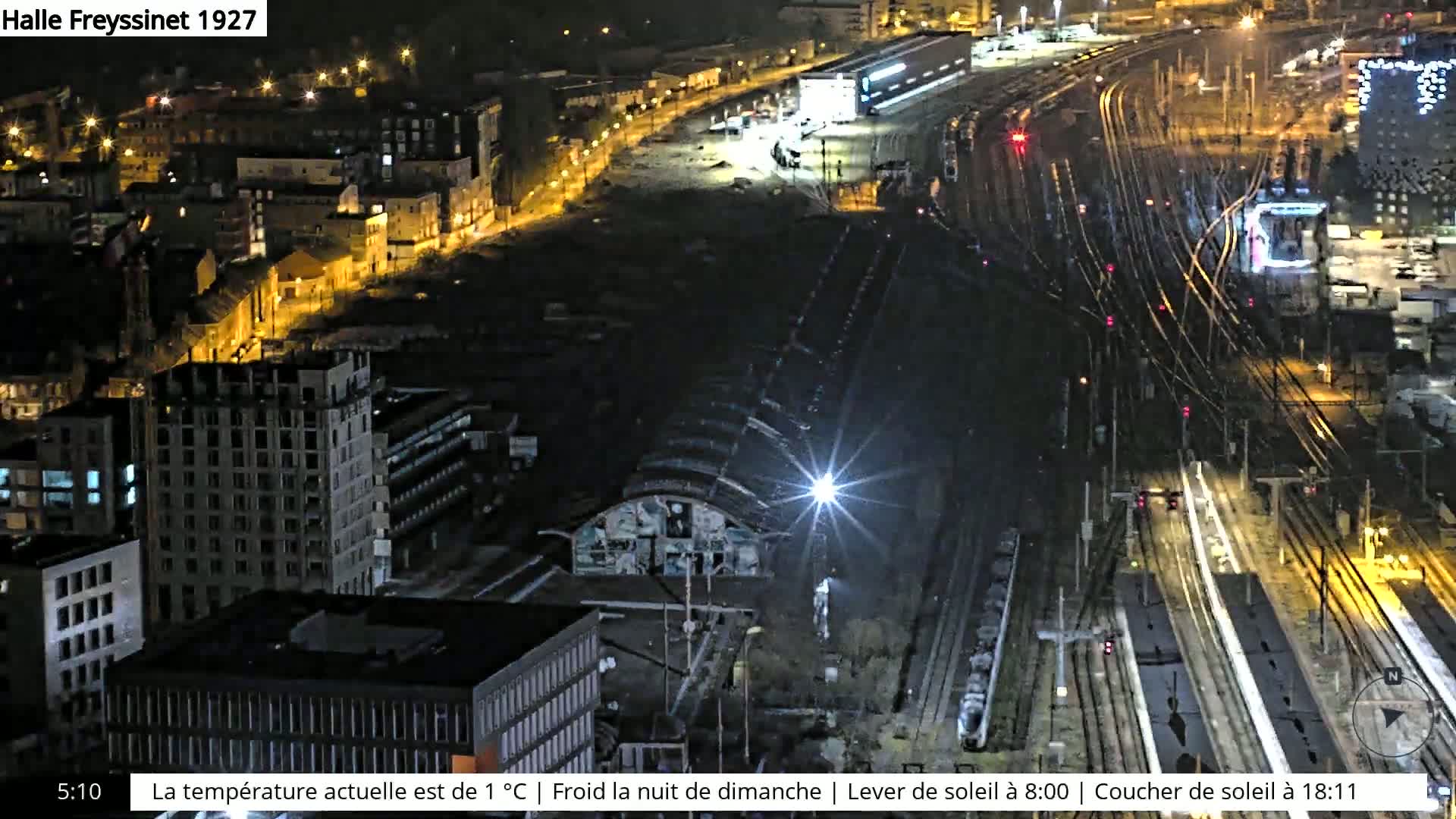 A nighttime aerial view of a train yard and surrounding city buildings, illuminated by streetlights and train lights, under a cold night sky.