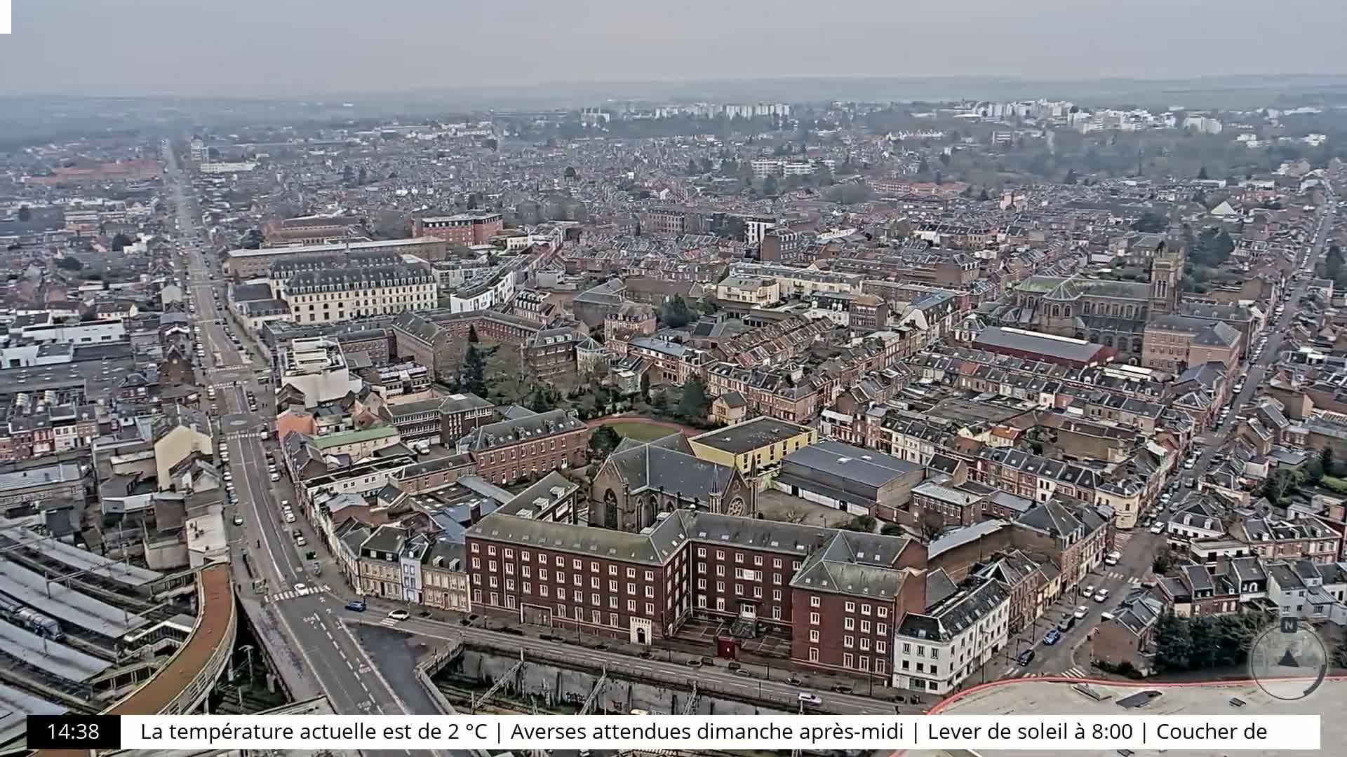 An aerial view of a city on an overcast day shows a dense collection of buildings and streets, with a major road and train tracks visible.