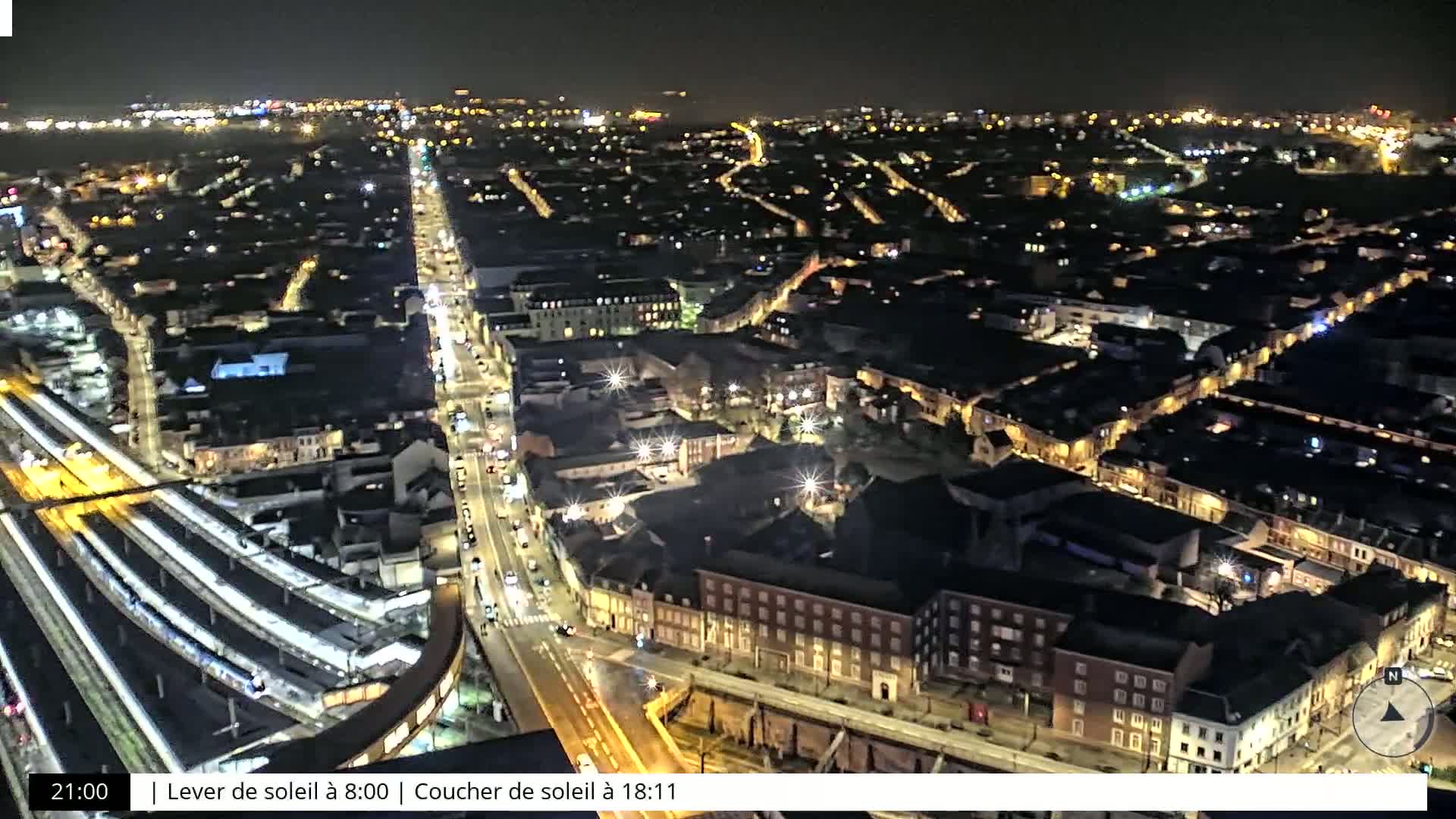 A nighttime, high-angle view of a city with illuminated streets and buildings, featuring a train station and surrounding roadways.