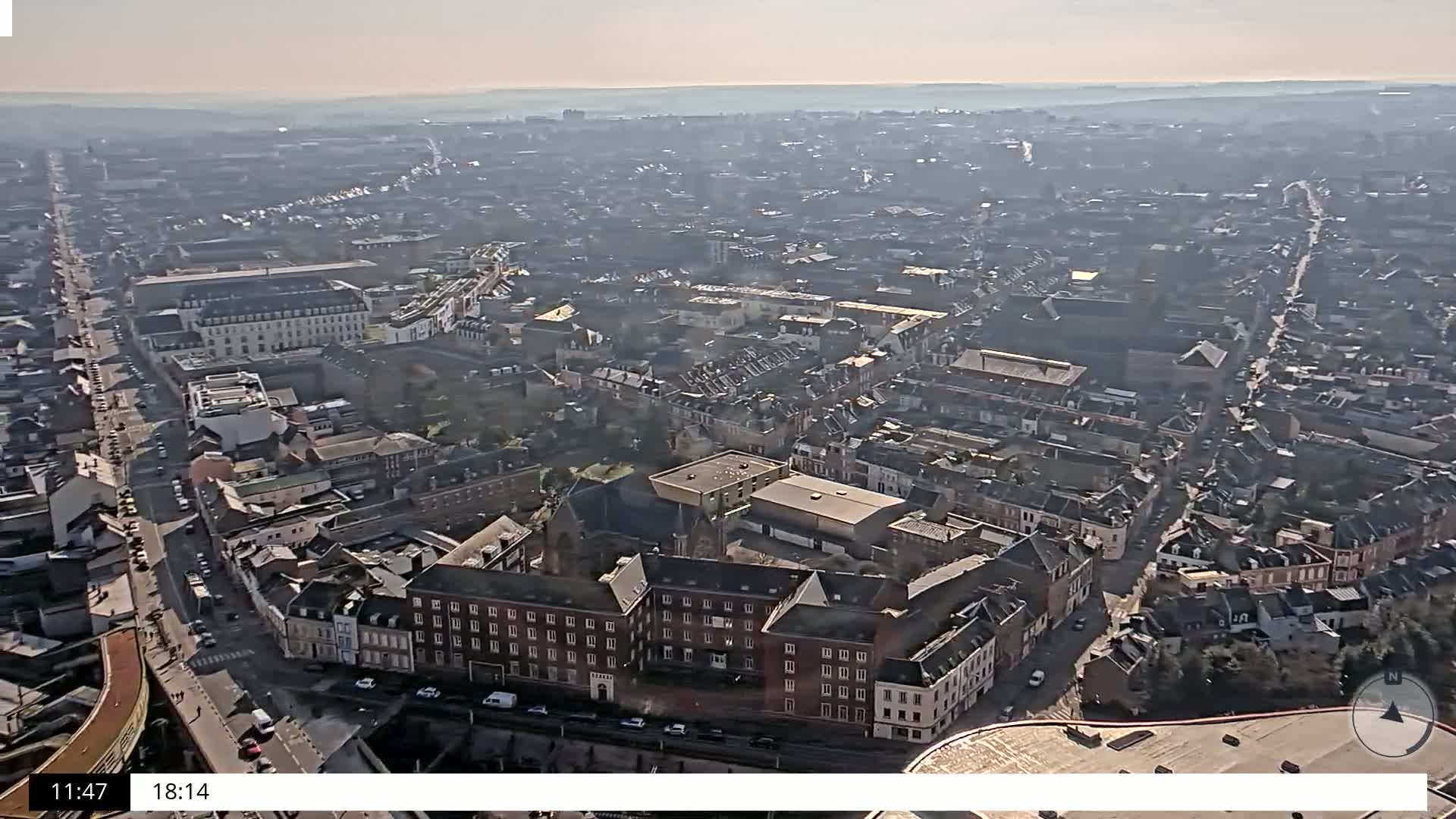 An aerial view shows a sprawling city on a hazy, sunny day, with many closely packed buildings and streets visible.
