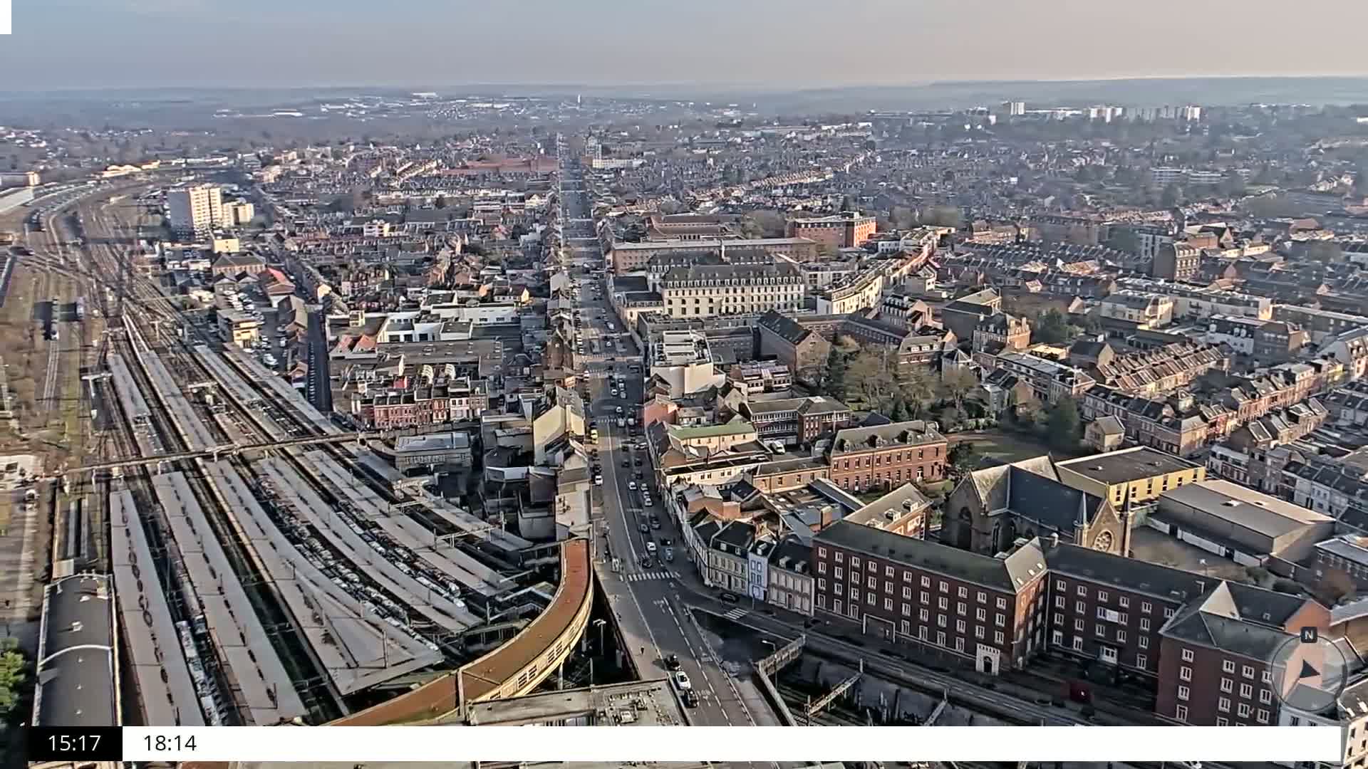 An aerial view shows a densely populated city with a large train station and many buildings under a clear, bright sky.