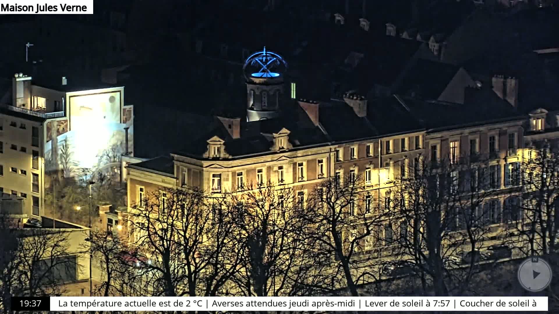 A nighttime view of a city block with several buildings illuminated, some bare trees in the foreground, and a building with a blue-lit dome.