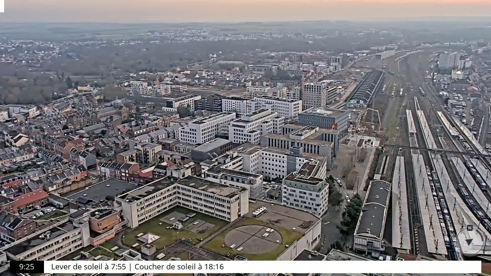 An aerial view of a city on a cloudy day shows a mix of residential and commercial buildings, a large train station with numerous tracks and trains, and a significant amount of undeveloped land.