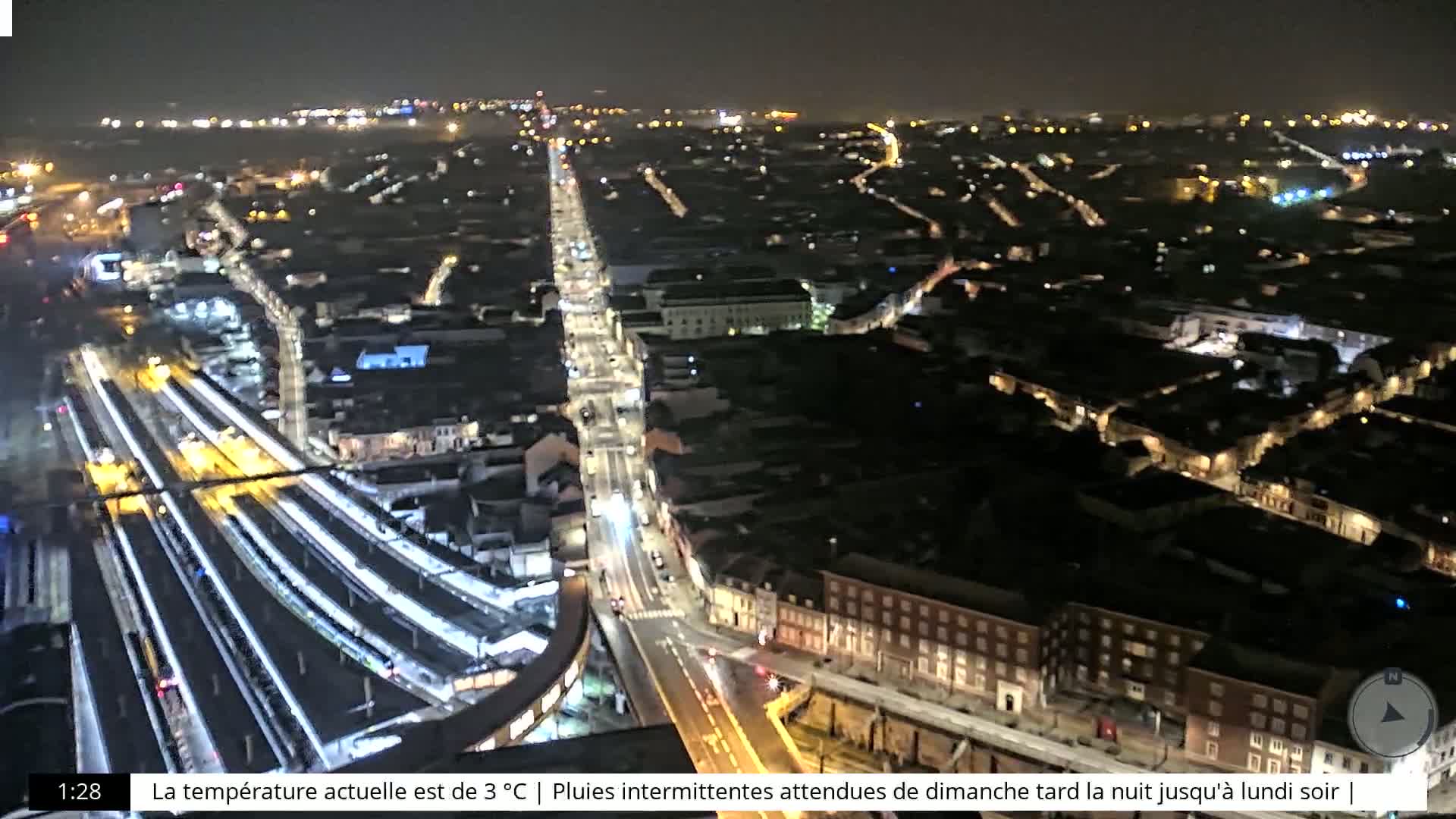A nighttime aerial view of a city with a train station and illuminated streets, under a clear sky.