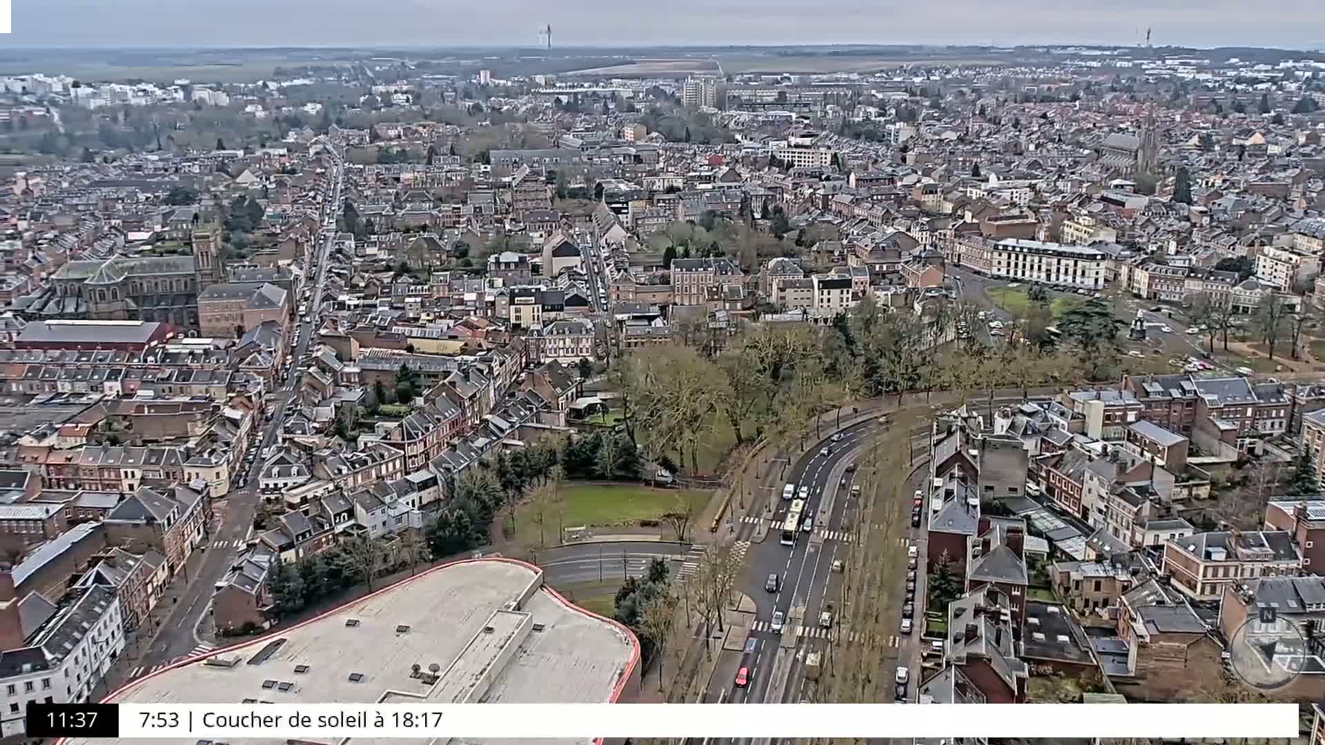 An aerial view shows a densely populated city under an overcast sky, with a road and park visible in the foreground.