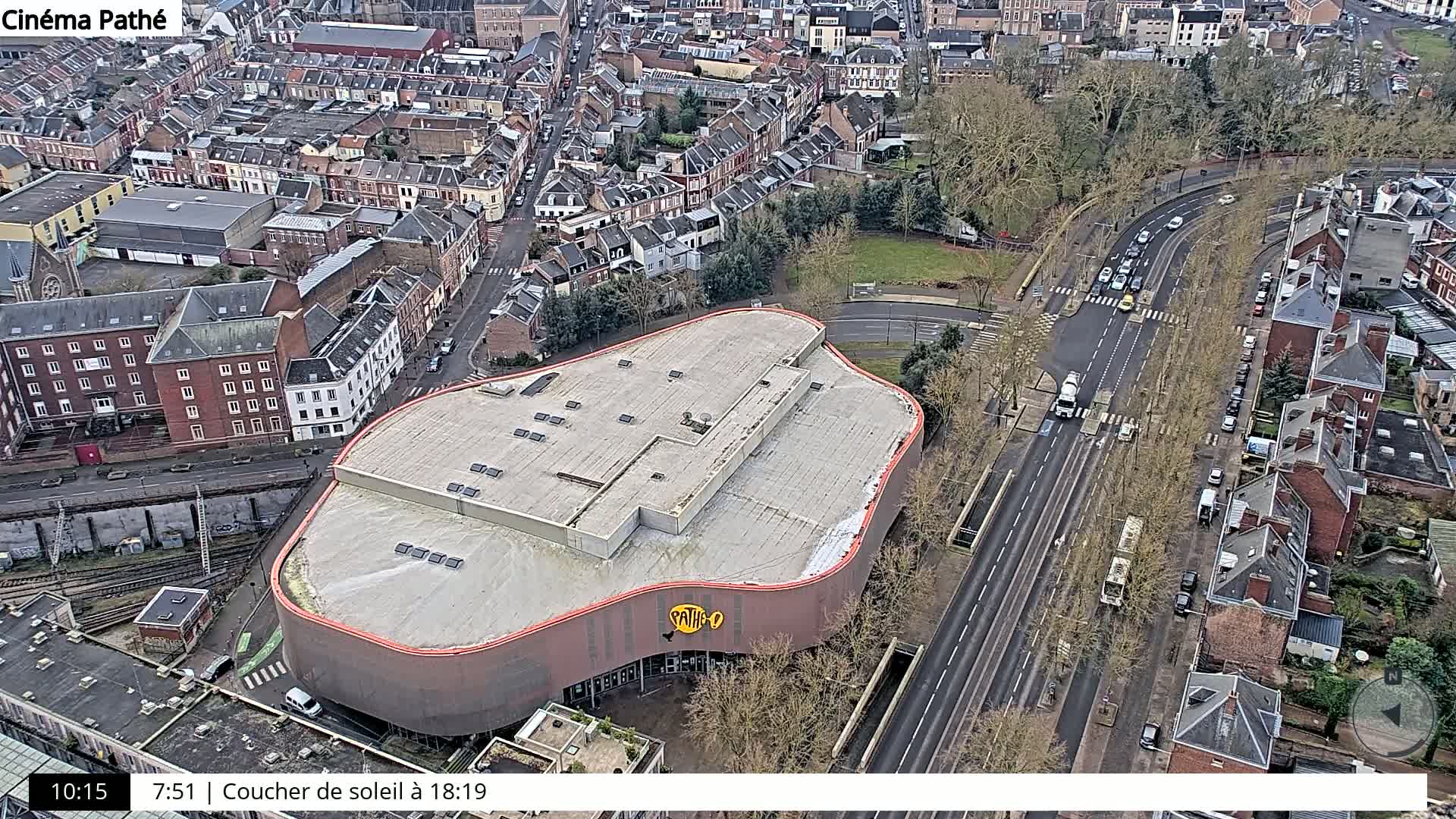 An aerial view shows a large, curved building with a flat roof next to a multi-lane road with sparse traffic, surrounded by a residential area with trees mostly bare of leaves.