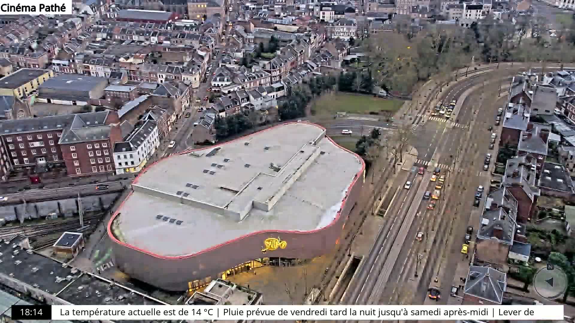 An aerial view shows a modern, curved building near a railway line and city streets with cars, under overcast skies.