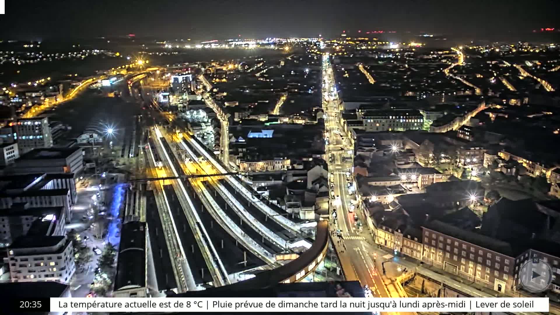 A nighttime aerial view of a city with brightly lit streets and a large train station, under a clear sky.
