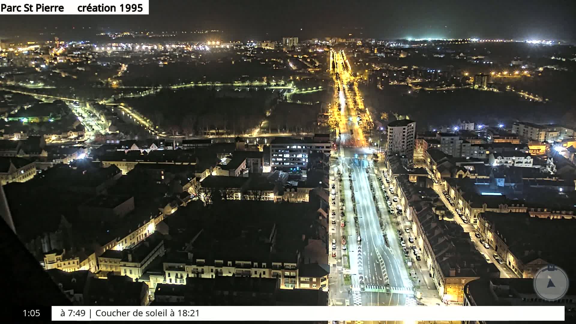 A nighttime aerial view shows a city with brightly lit streets and buildings, surrounded by darker areas of trees and parkland.