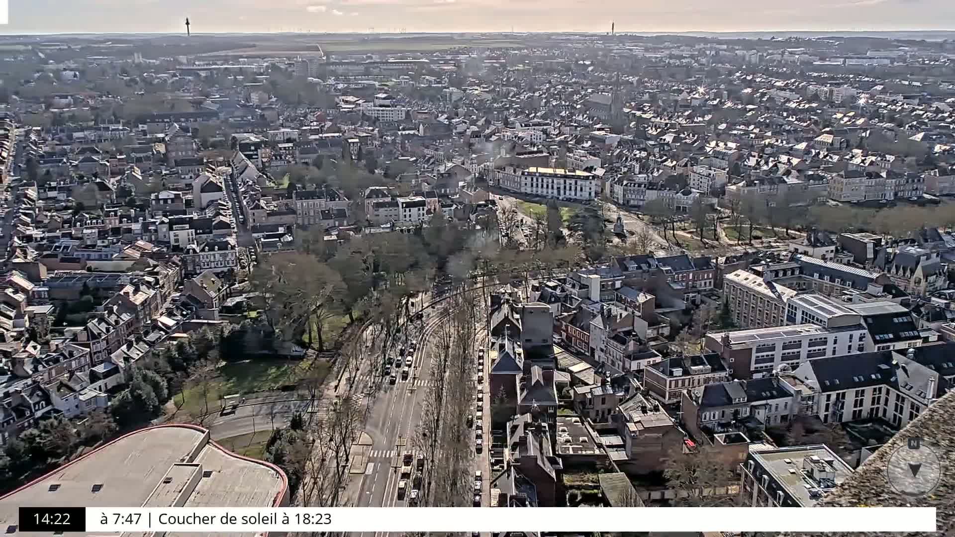 A high-angle, long shot of a city on a sunny day shows many residential buildings and a long road with parked cars.