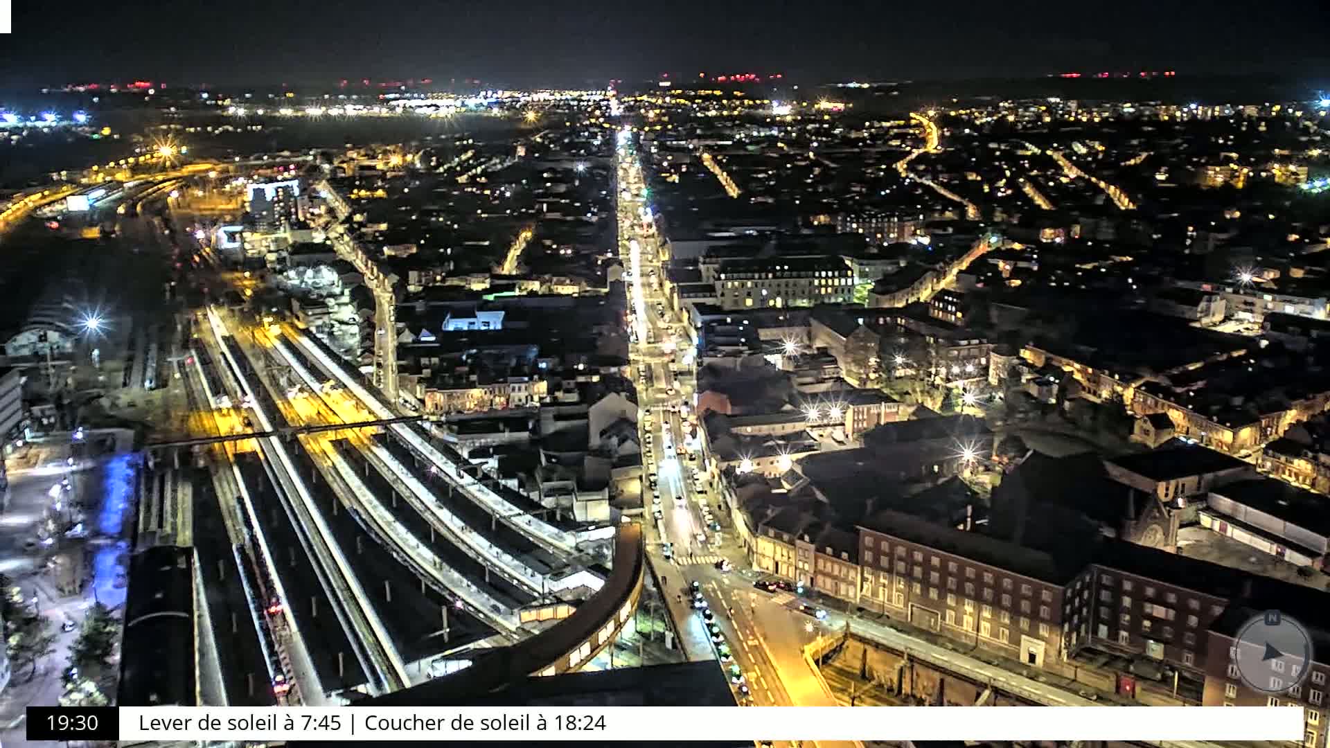 A nighttime aerial view of a city with brightly lit streets and a train station, under a clear sky.