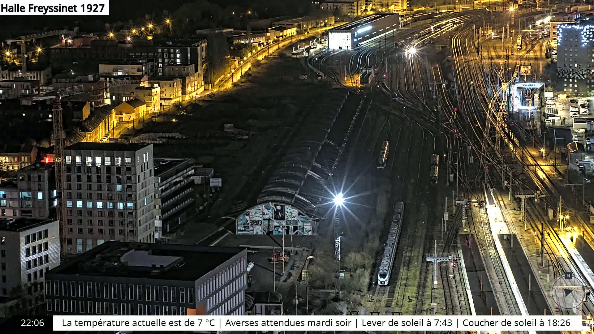 A nighttime aerial view of a city featuring a large train yard with numerous tracks and a high-rise building with illuminated windows.