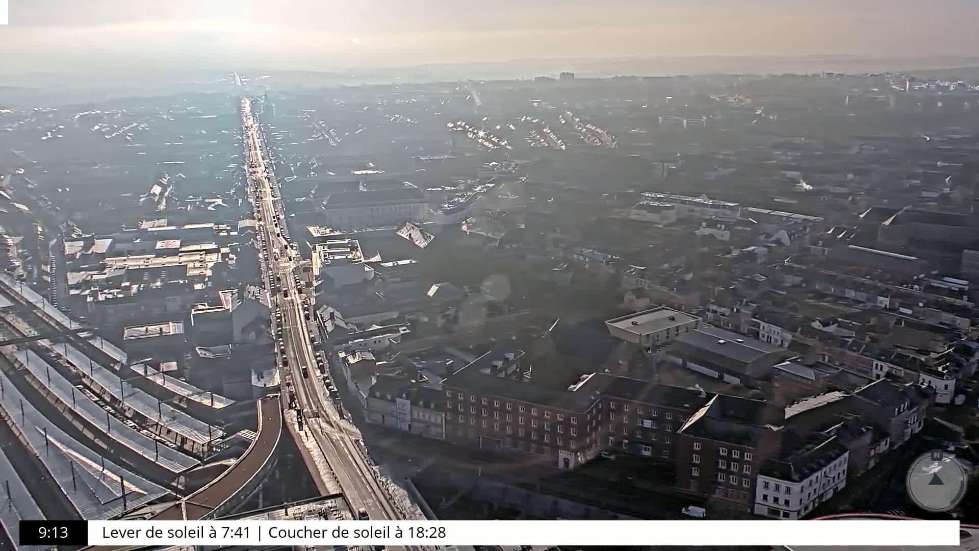 A hazy, high-angle view of a city with a long road and a train station visible, on a somewhat bright day.