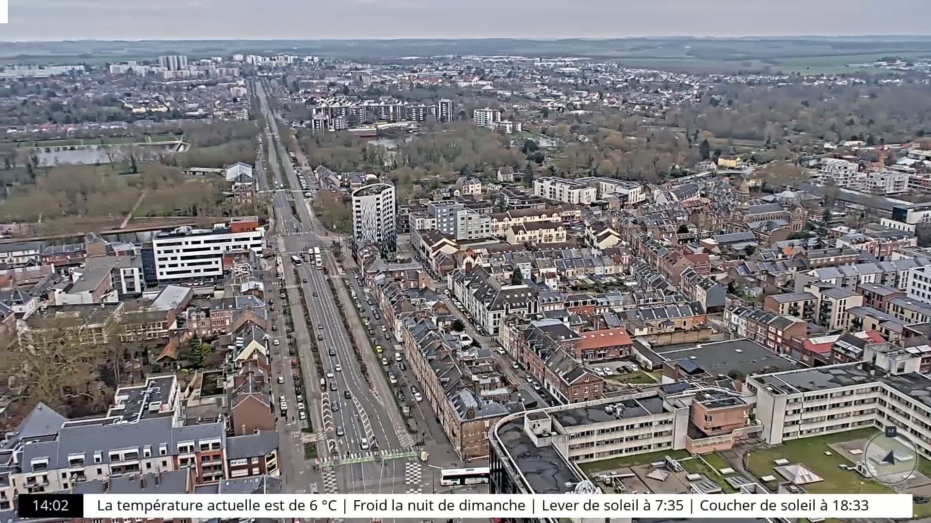 An aerial view of a city on an overcast day shows a long road lined with cars, surrounded by residential buildings and green spaces.