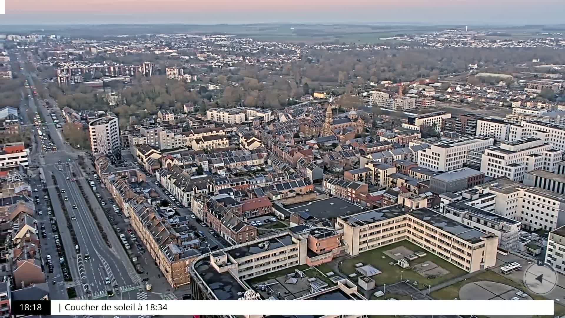 An aerial view of a densely populated city under a twilight sky shows a mix of residential buildings and larger structures, with a long road cutting through the center.