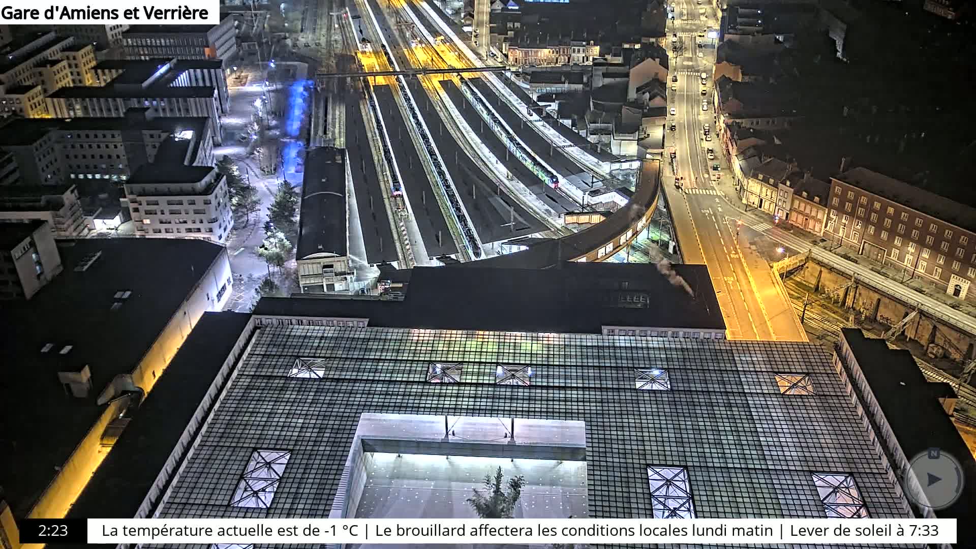 A nighttime, high-angle view of a train station and surrounding city, illuminated by artificial lights.