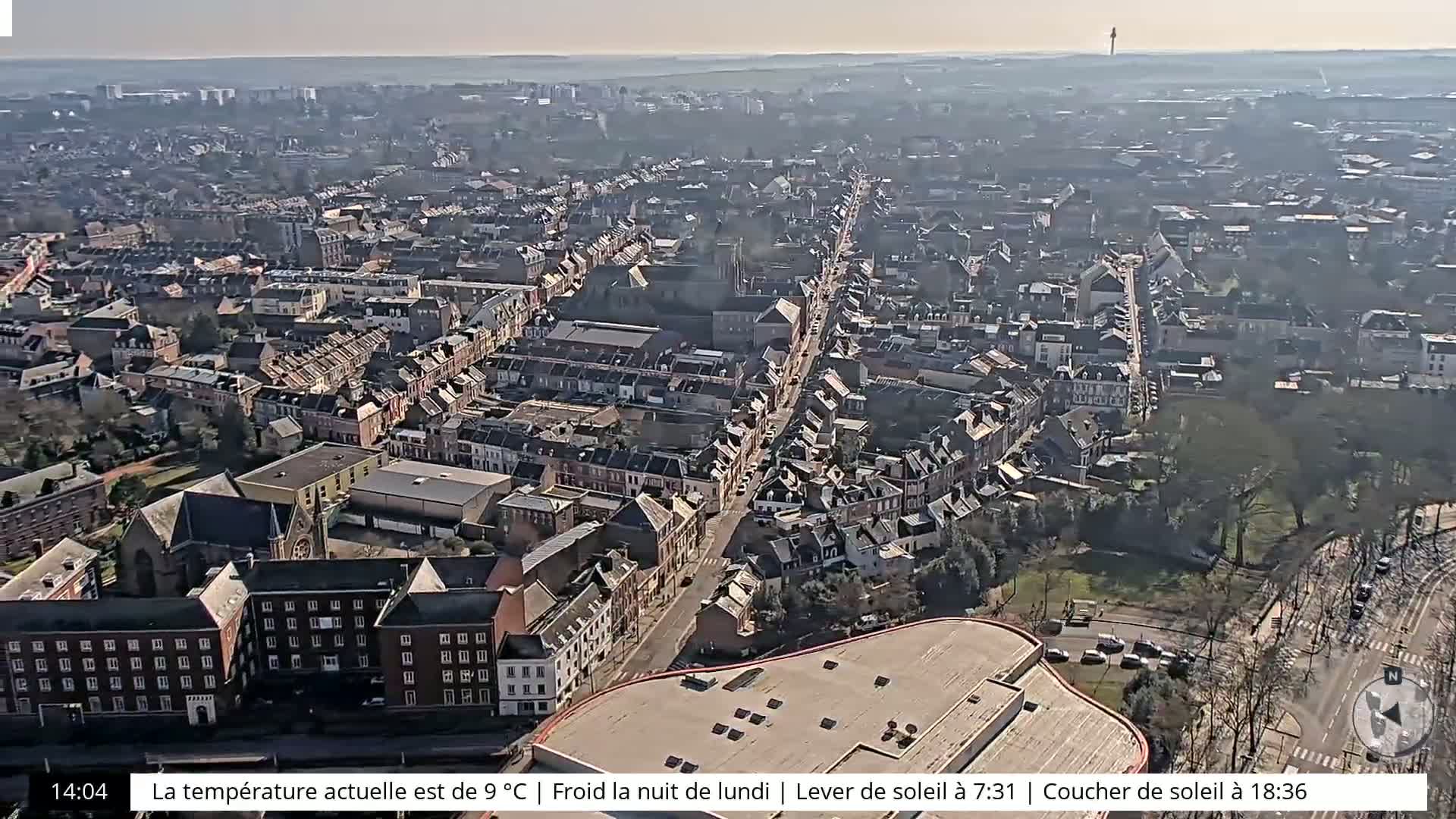 An aerial view of a city on a clear, slightly hazy day shows a long street lined with buildings, and a large parking lot.