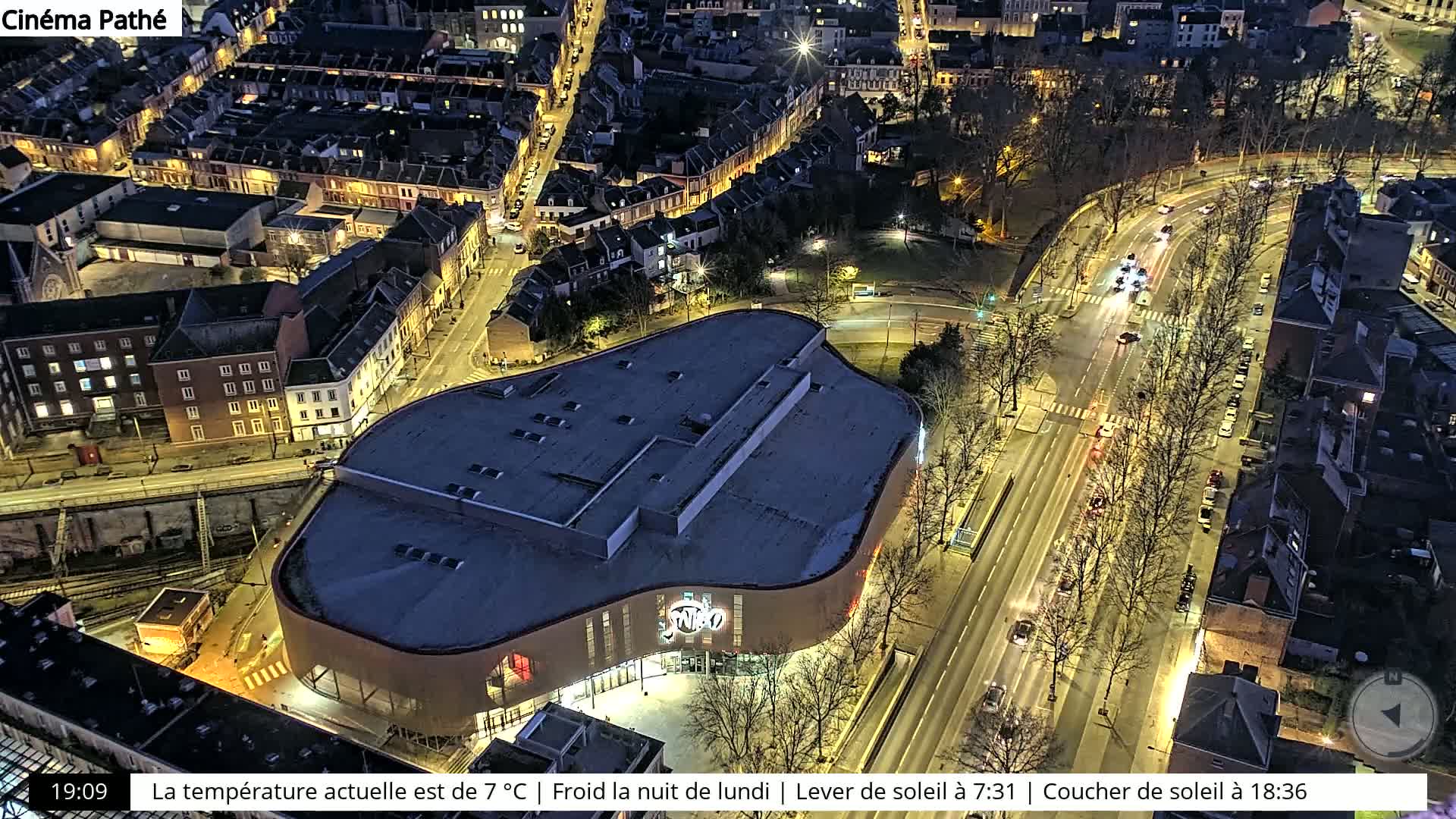 An aerial nighttime view of a city with a large, curved building illuminated, roads with cars, and surrounding buildings, under a cold night sky.