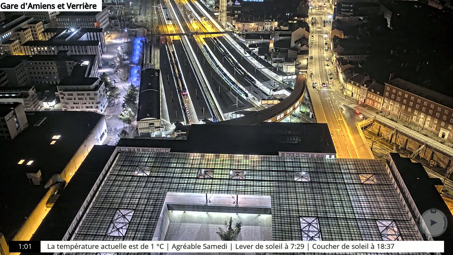 A nighttime aerial view of a train station and surrounding city streets, illuminated by artificial lights.