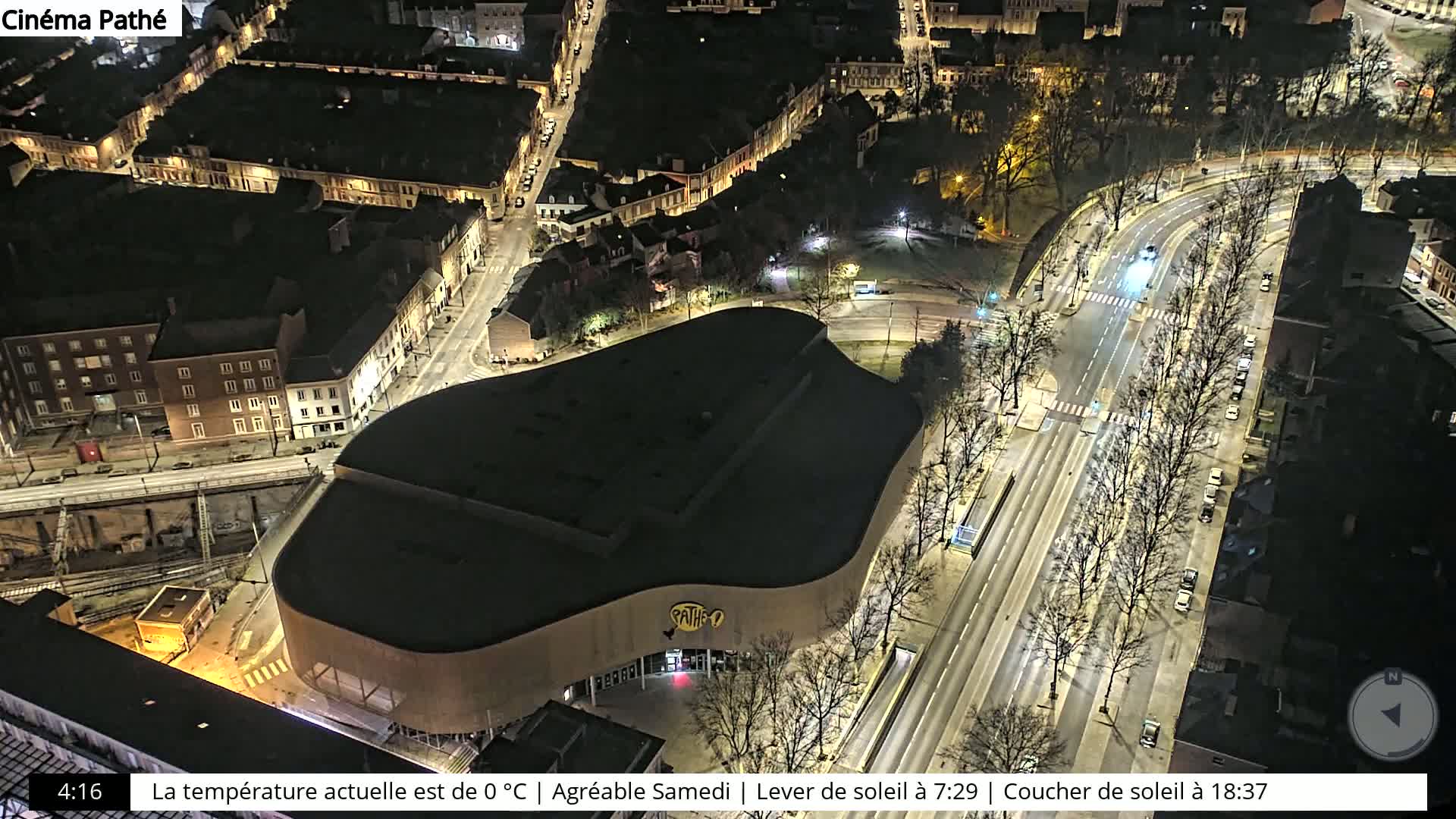 An aerial nighttime view of a city street with a large, dark building, possibly a cinema, situated beside it, surrounded by other buildings and bare trees.