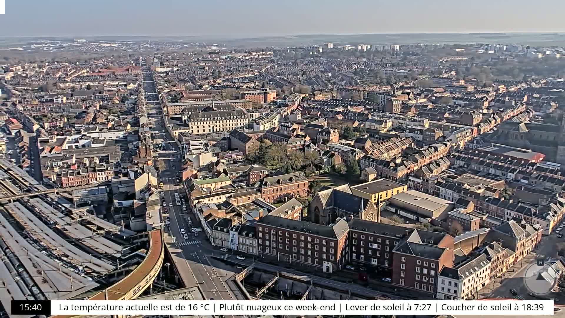 A high-angle, long shot of a city on a partly cloudy day, showing a long road lined with buildings and a train station.
