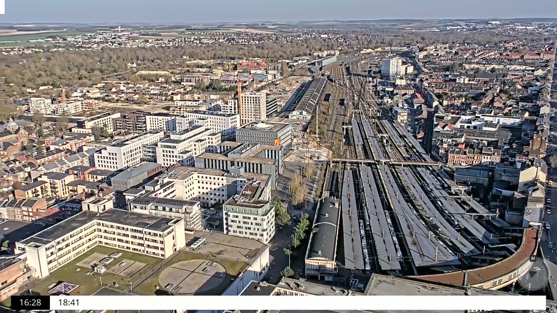 An aerial view of a city on a sunny day shows a large train station surrounded by modern and older buildings, with undeveloped land visible in the distance.
