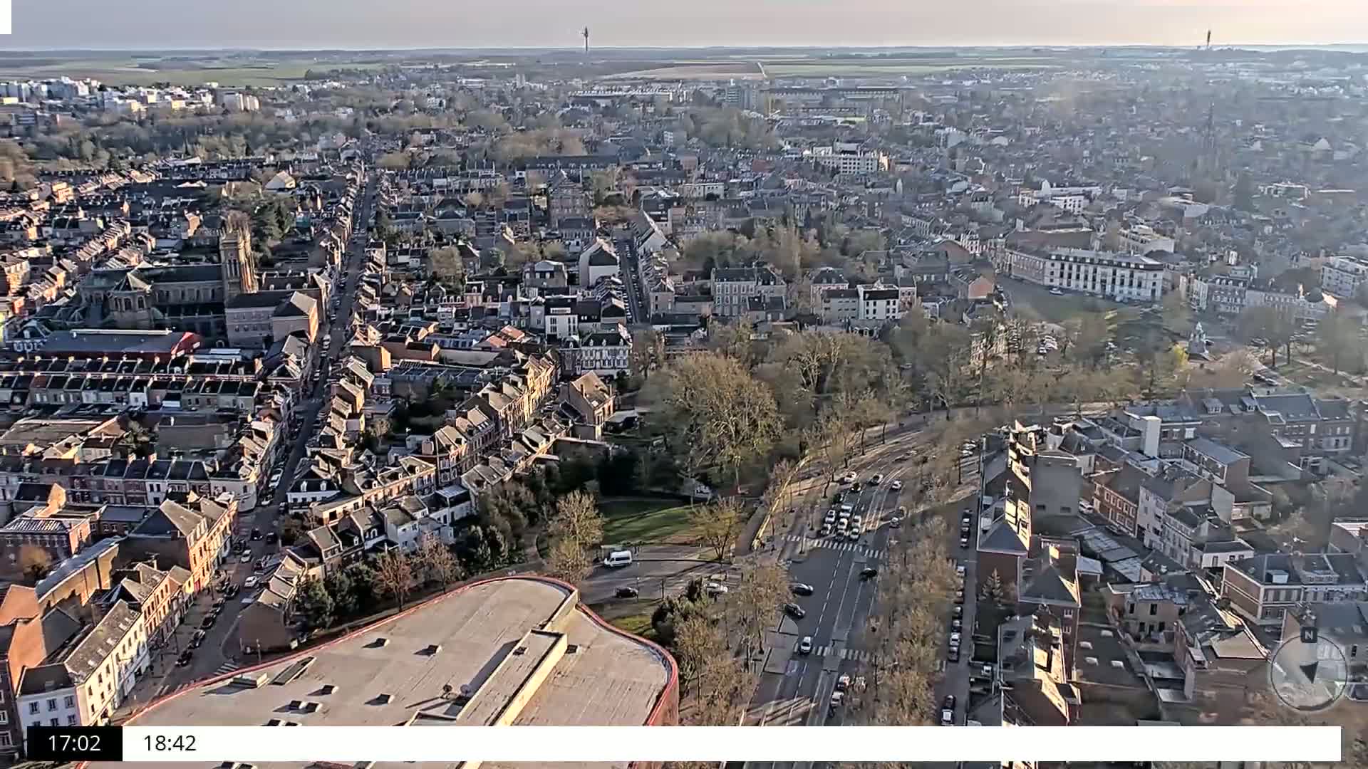 An aerial view of a densely populated city on a partly sunny day shows many buildings and streets, with some trees and a large parking lot visible.